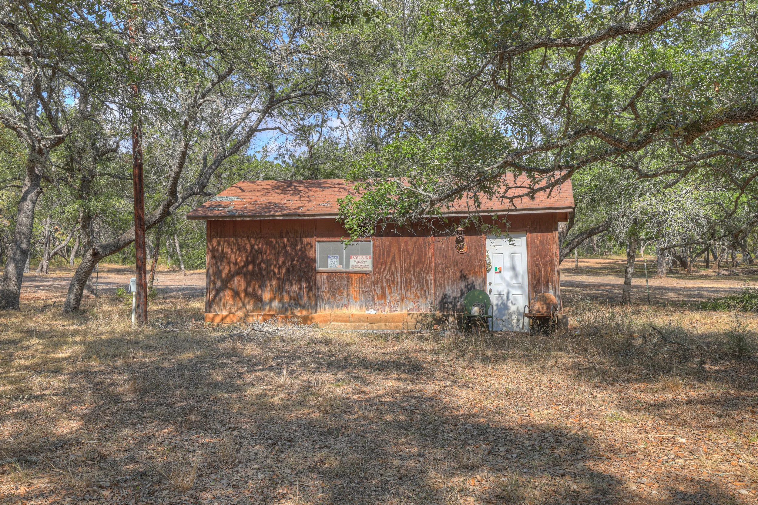 A small, weathered wooden building with a white door and a window, surrounded by trees and dry grass, with shadows from the branches crossing the front of the building.