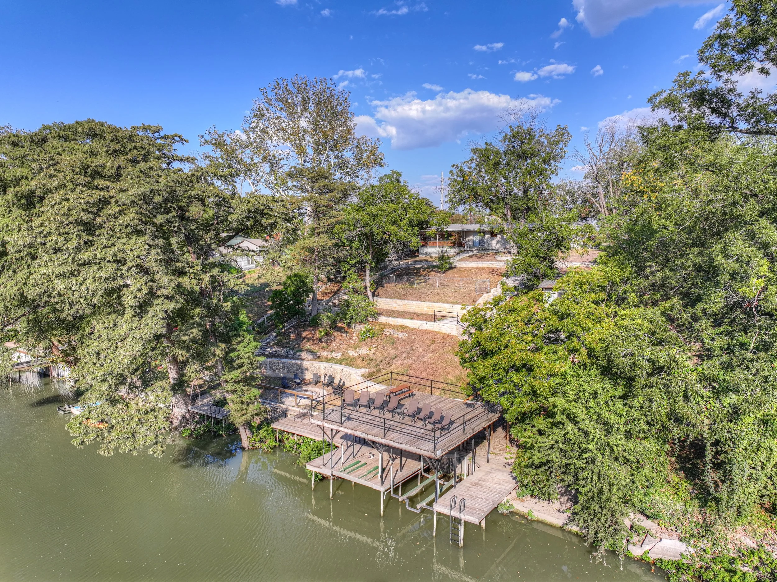 A lakeside backyard with a wooden deck and patio furniture, surrounded by trees and overlooking a green water body on a sunny day.