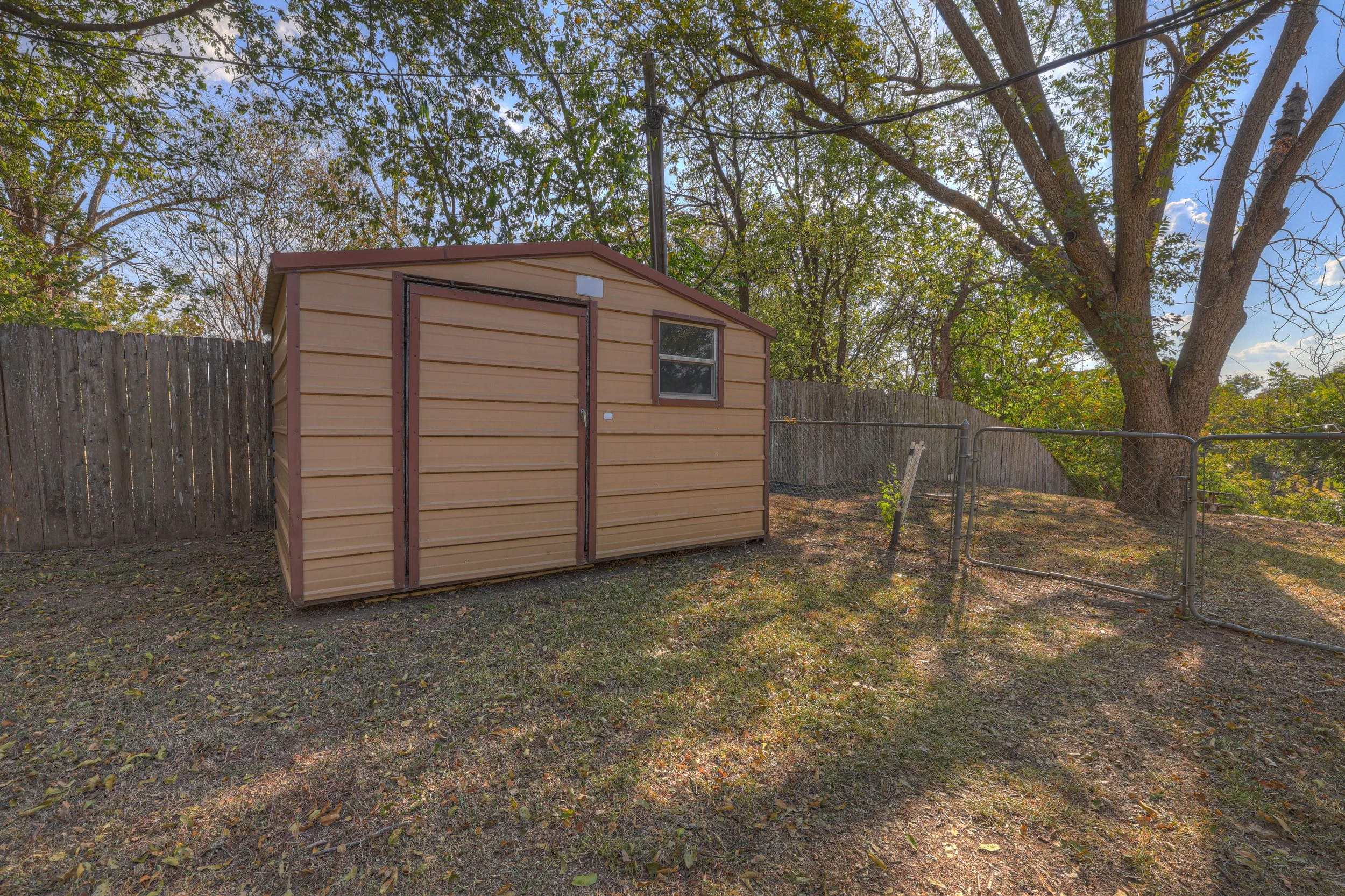 A backyard with a small tan metal shed, a chain-link fence, a large tree, and a wooden privacy fence, under a bright blue sky with scattered clouds.
