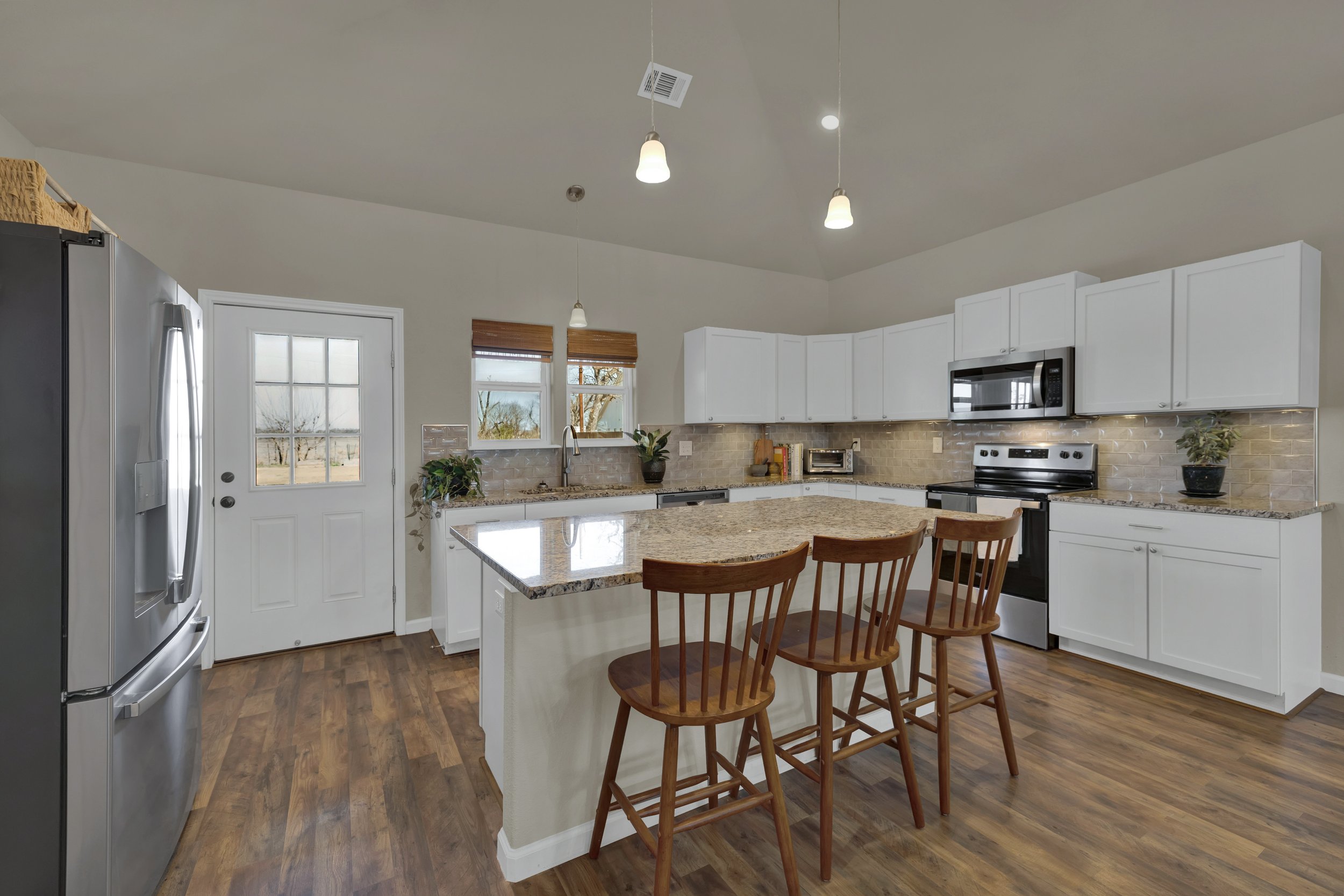 Kitchen with white cabinets, granite countertops, stainless steel appliances including a refrigerator, oven, microwave, and toaster. There are three wooden bar stools at a breakfast bar, a window with blinds, and plants on the counter.