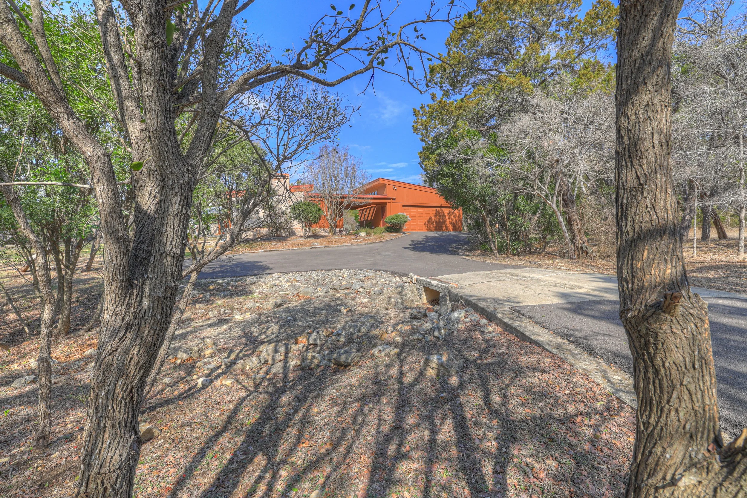 Residential driveway leading to a brown house, surrounded by trees with some bare branches, under a blue sky