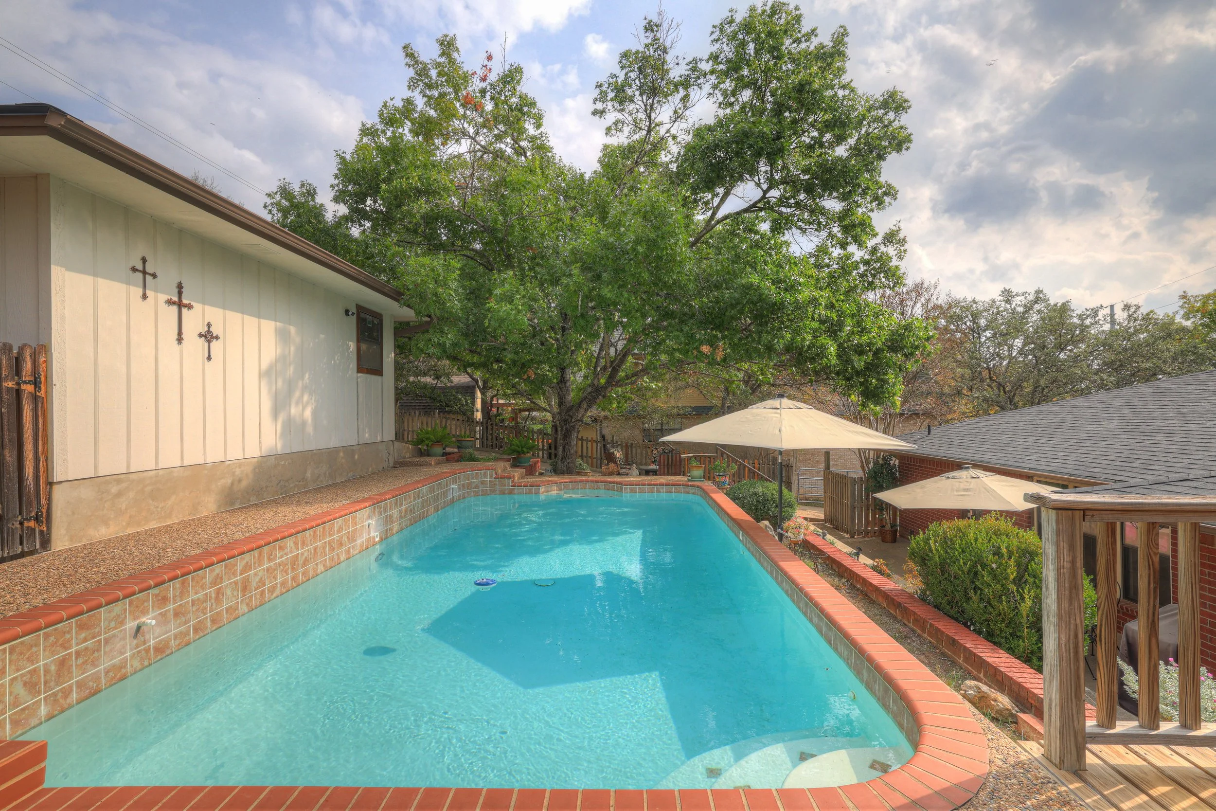A backyard with a swimming pool, large trees, umbrellas, and a house with a gray roof and a screened porch. The pool is surrounded by brick and tile, with a wooden fence and garden in the background.