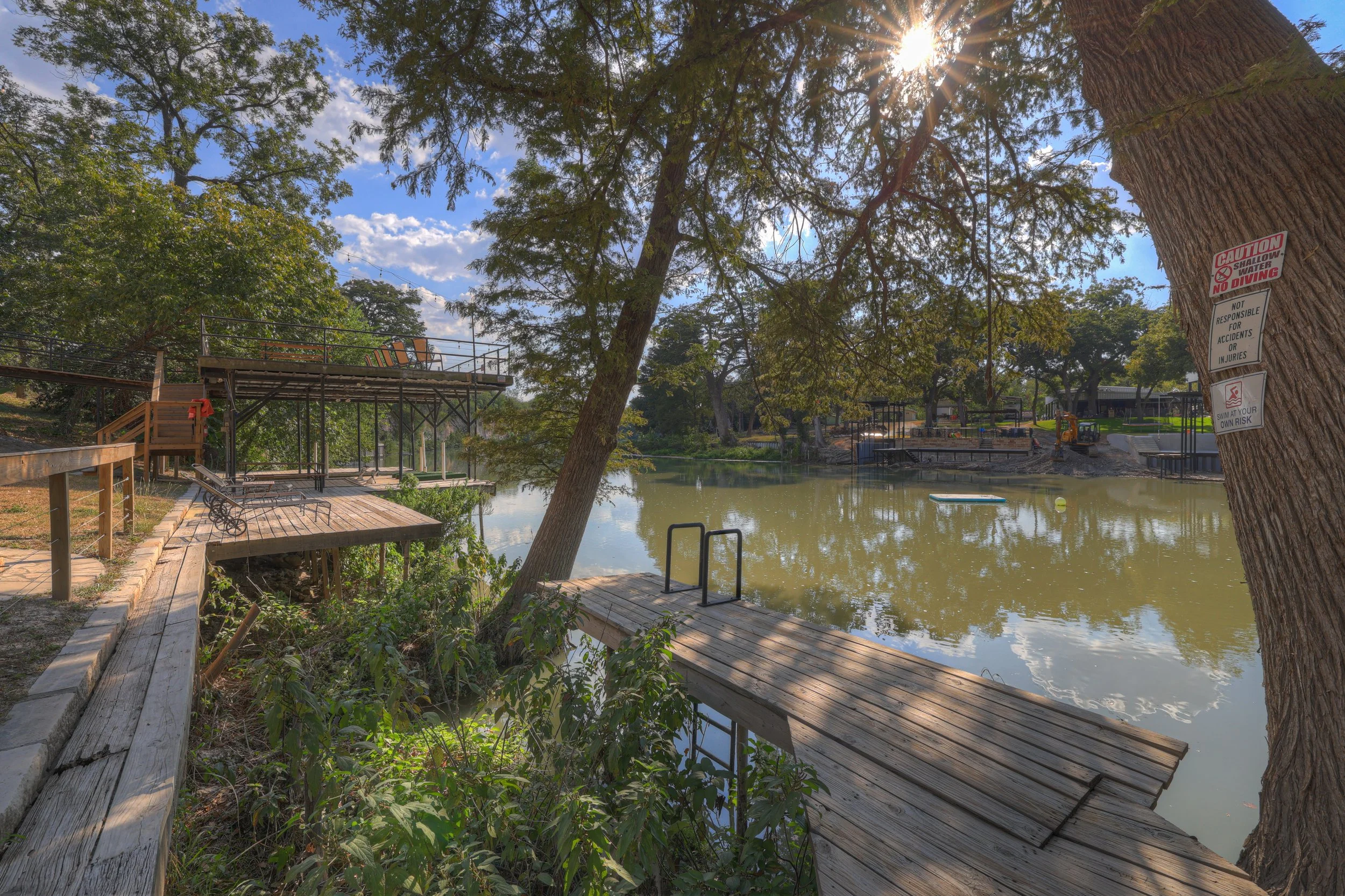 A lakeside park with a wooden dock, trees, and a water slide in the background on a sunny day with some clouds.