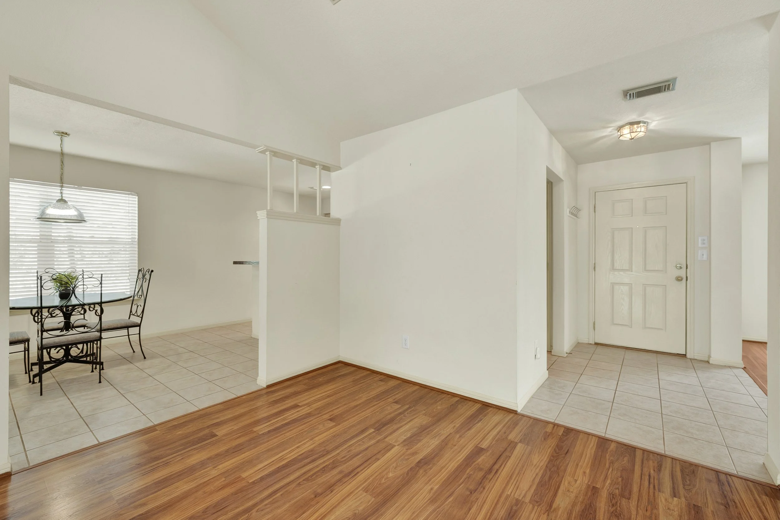 Empty living room with hardwood flooring, beige tiled entryway, white walls, and a white front door. A dining area with a black metal table and chairs is visible to the left, with a window with blinds and a hanging light fixture above.