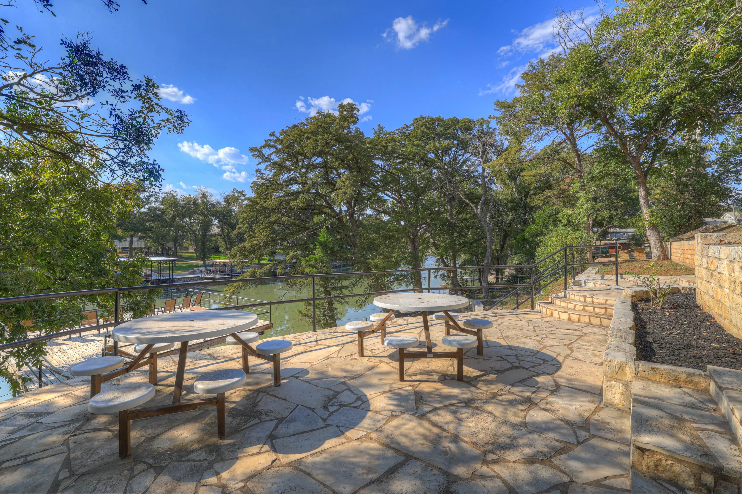 An outdoor patio with two round tables and attached stools, overlooking a river with trees and a clear blue sky.