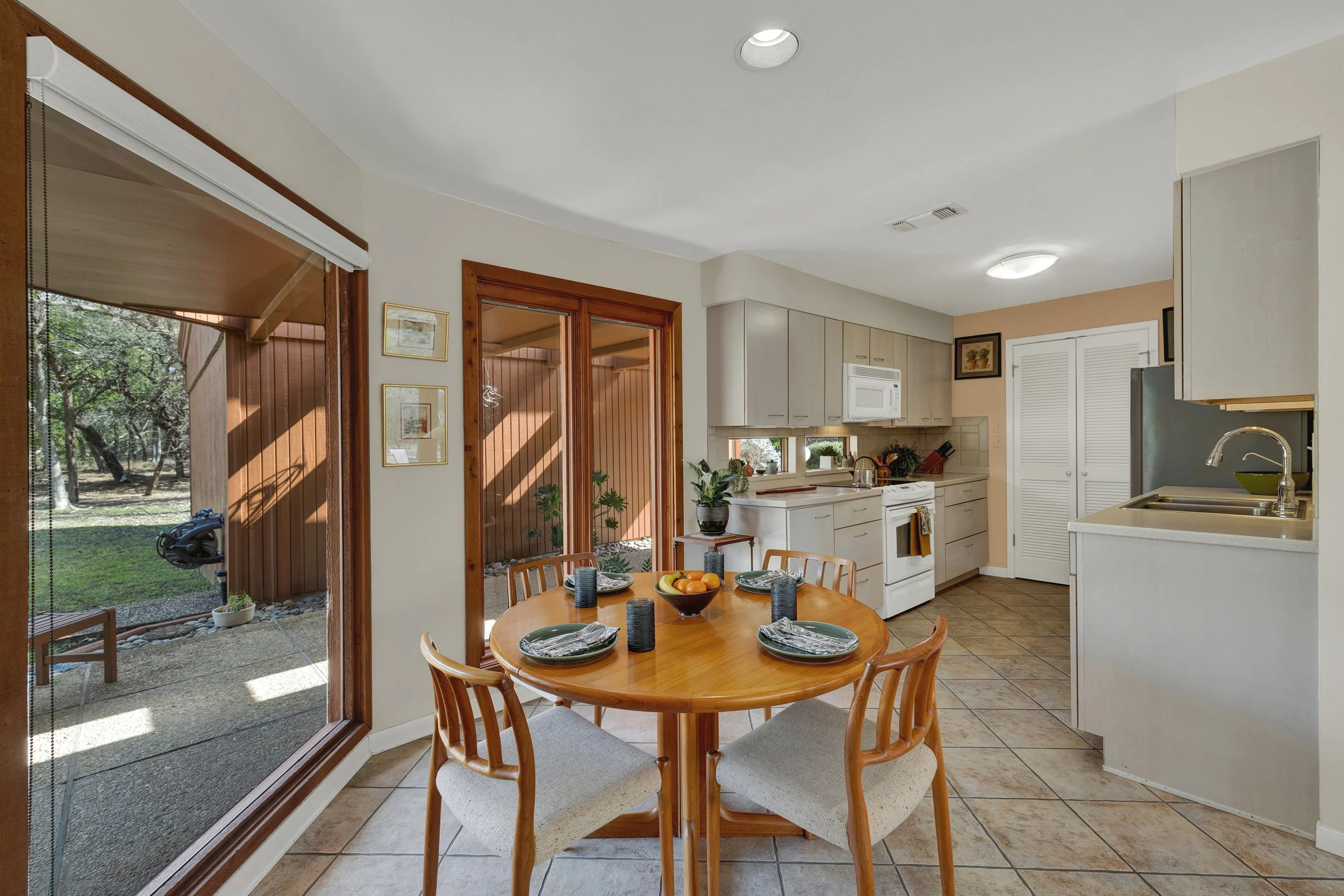 Kitchen and dining area with a round wooden table set with plates and glasses, surrounded by four chairs. The space features a sliding glass door to the outside yard, white cabinets, and a window over the counter.
