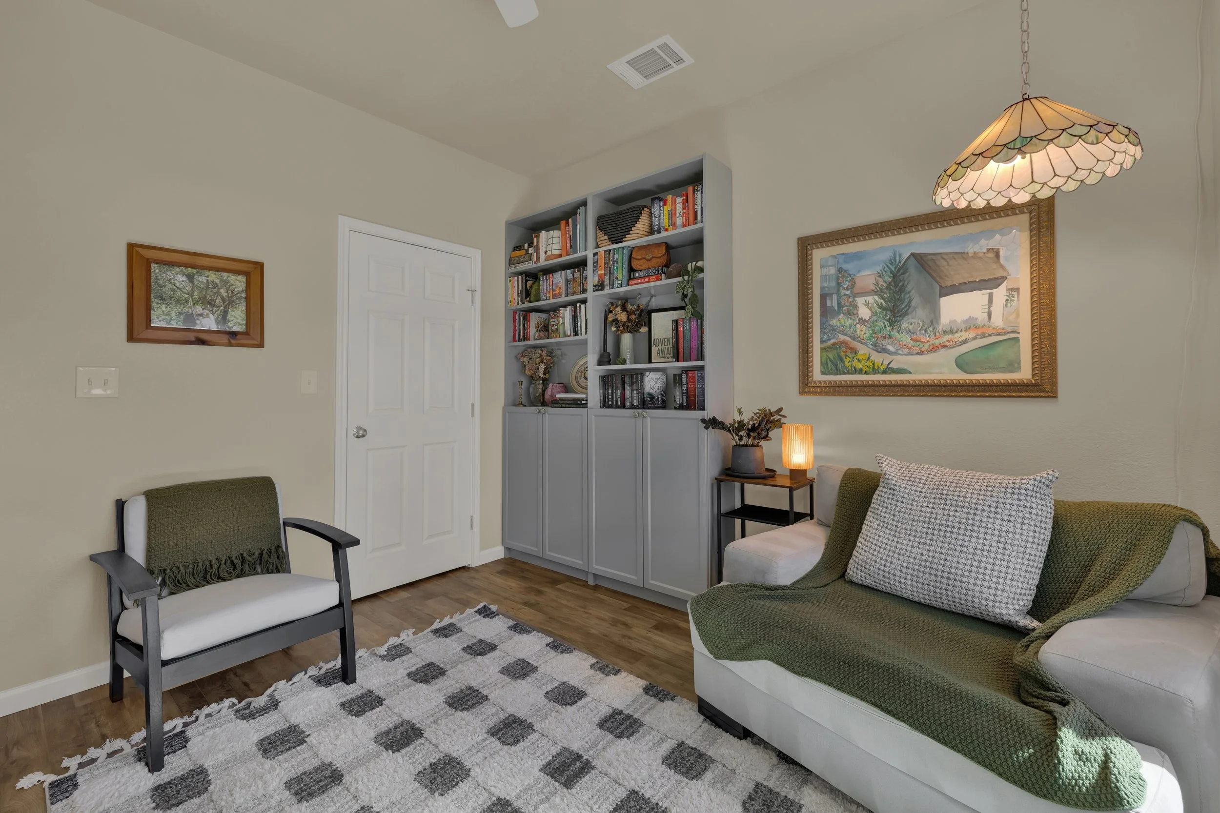 Living room with a green and white sofa, a black side table with a lamp, a patterned rug, a bookshelf, and wall art.