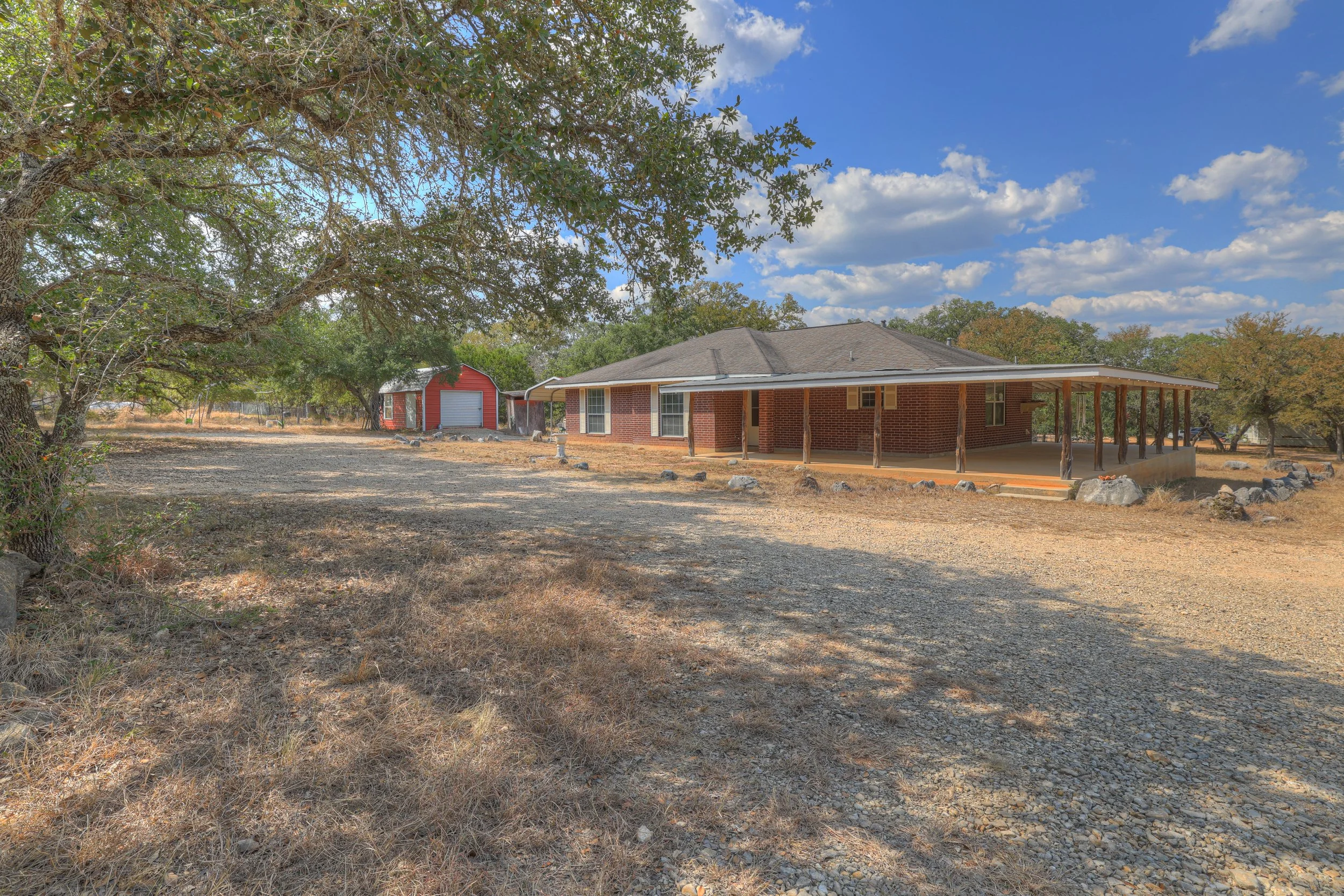A brick house with a large covered porch situated on a spacious, dry, and rocky yard with some trees and a red shed in the background under a partly cloudy sky.
