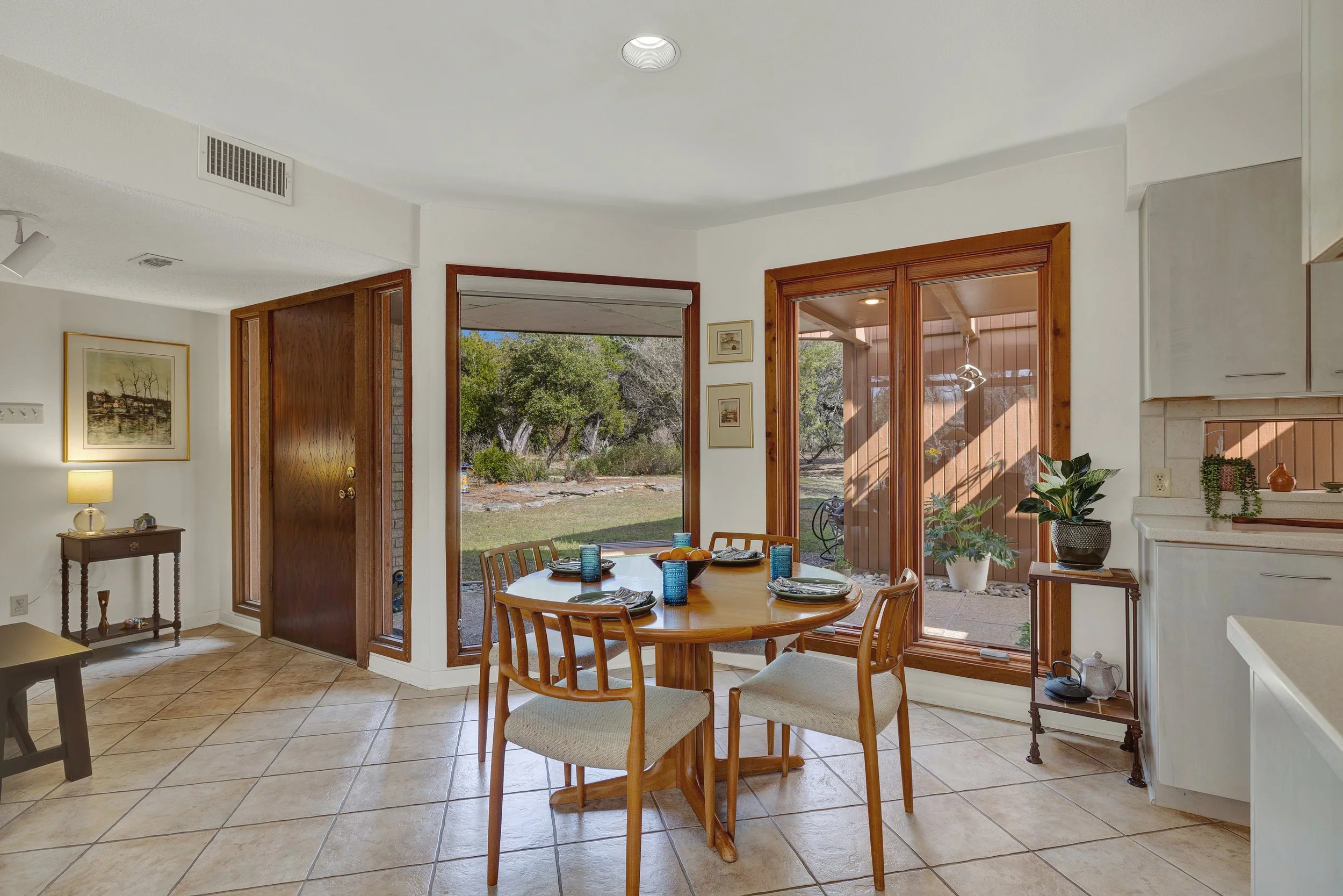Dining area with a round wooden table, four chairs, and large sliding glass doors leading to a backyard with trees and shrubs.