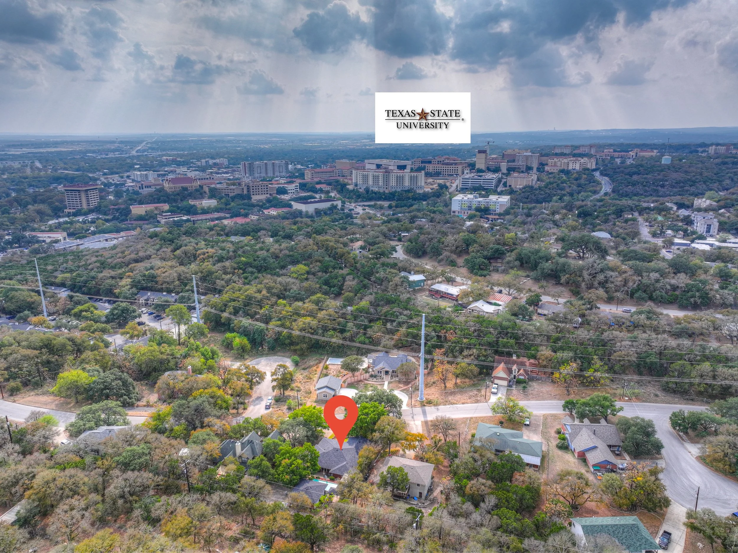 Aerial view of a city with a mix of residential houses, trees, and taller buildings in the background. A red location pin marks a house in the lower center. The sky is partly cloudy with sun rays breaking through.