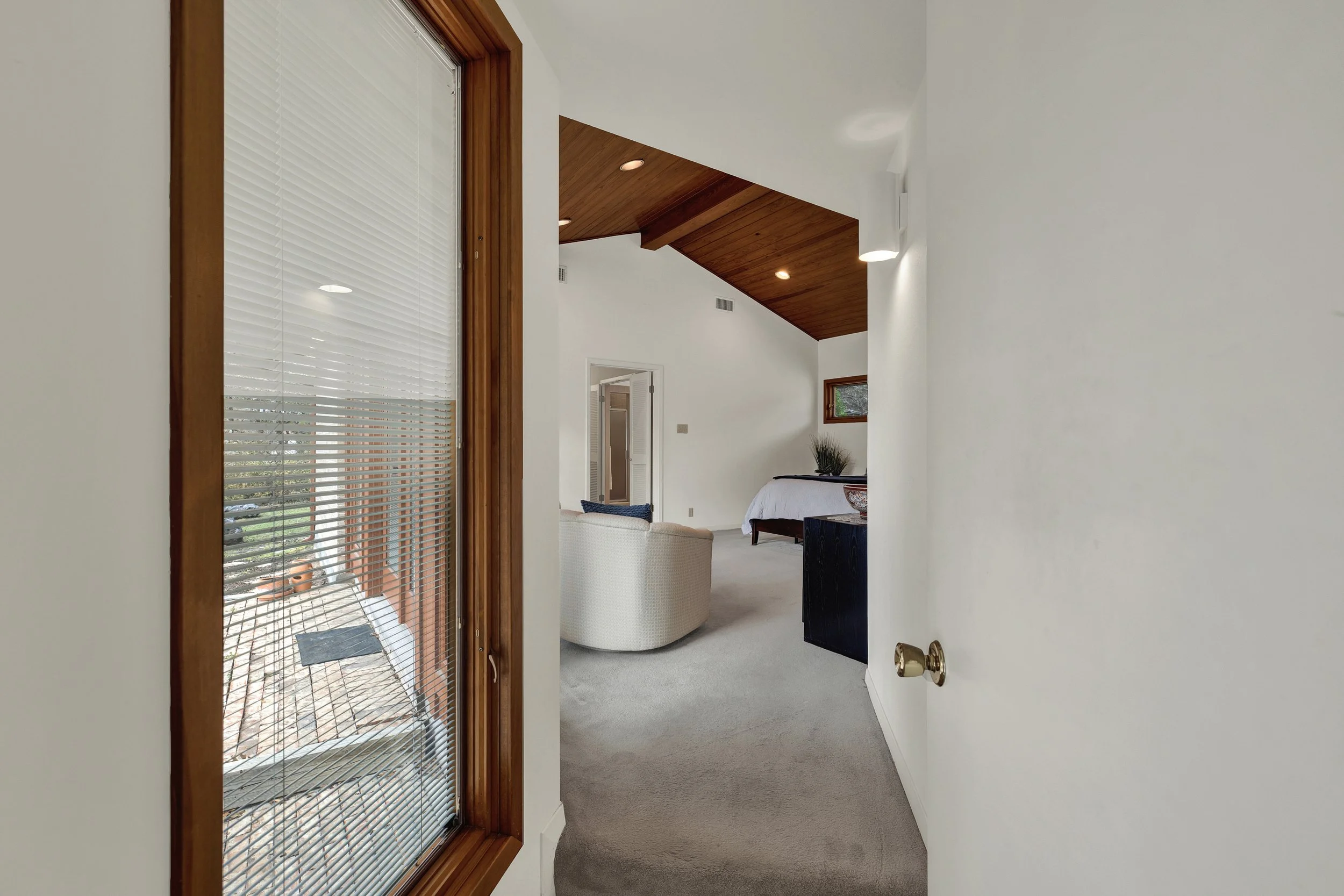 Interior view of a living room with white walls, a vaulted wooden ceiling, and a window with blinds showing an outdoor patio.