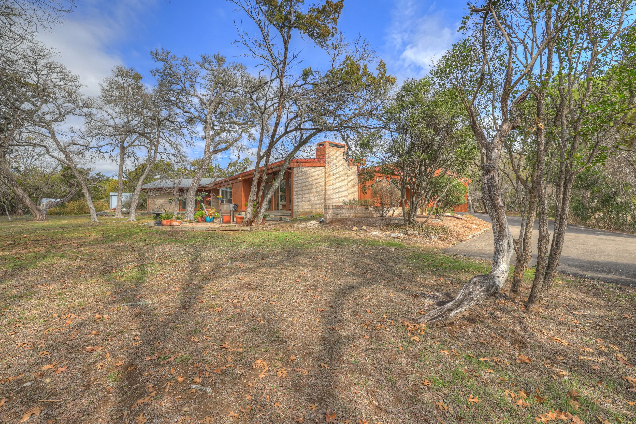 A mid-century modern house with brick and wood exterior in a yard with leafless trees, some potted plants, and an asphalt driveway, under a partly cloudy sky.