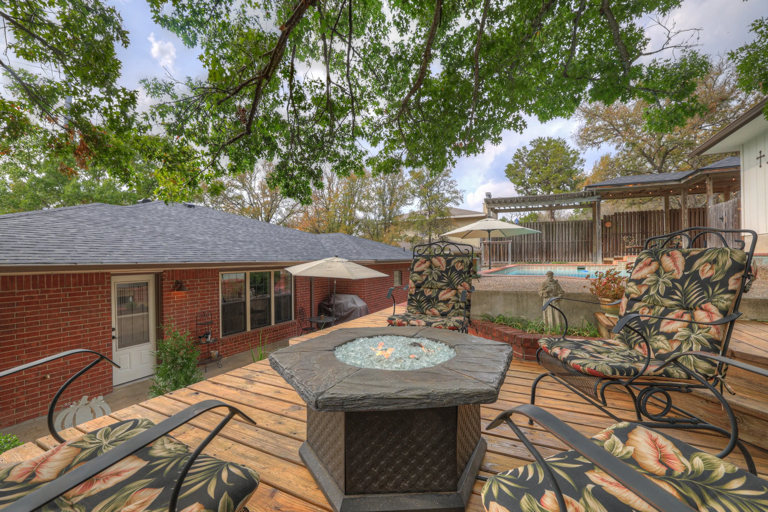 Backyard patio with seating area, fire pit, umbrellas, pool, and surrounded by trees and a brick house.