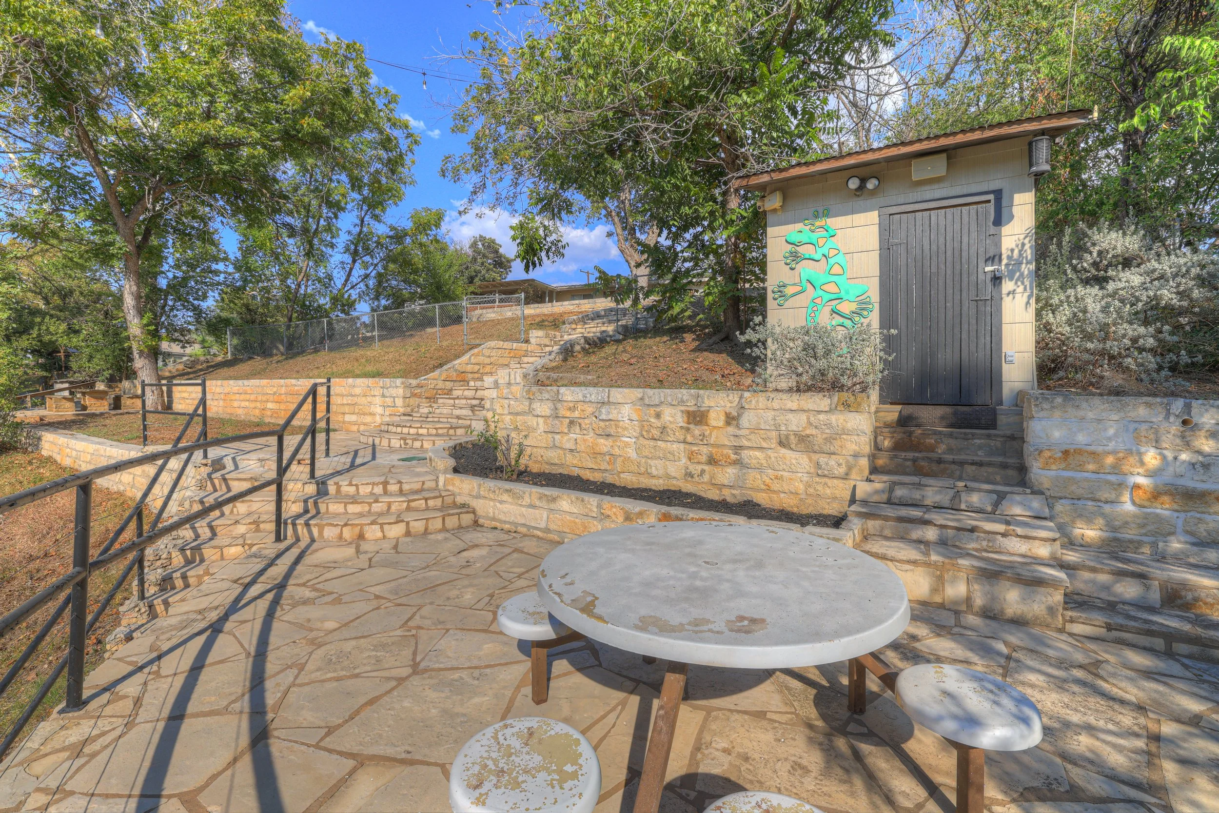 Outdoor patio with stone flooring, black metal railing, wooden stairs, a small gray shed with a painted green gecko on the door, surrounded by trees and foliage, under a partly cloudy sky.