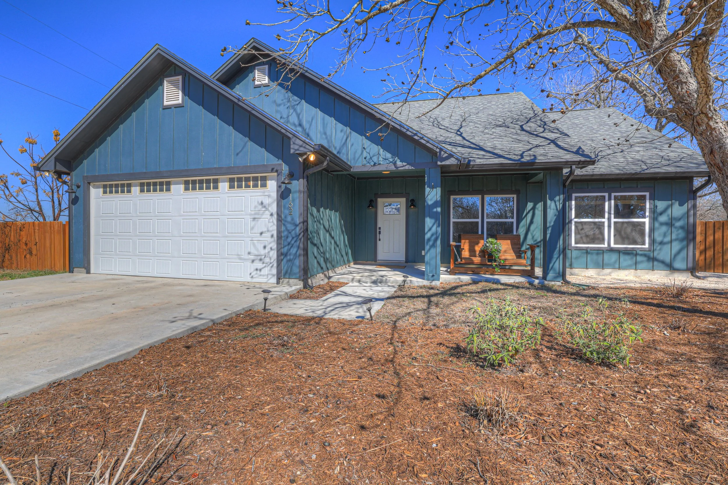 Front view of a blue house with a white garage door, a porch with a wooden bench, and leafless trees in the yard during daytime.