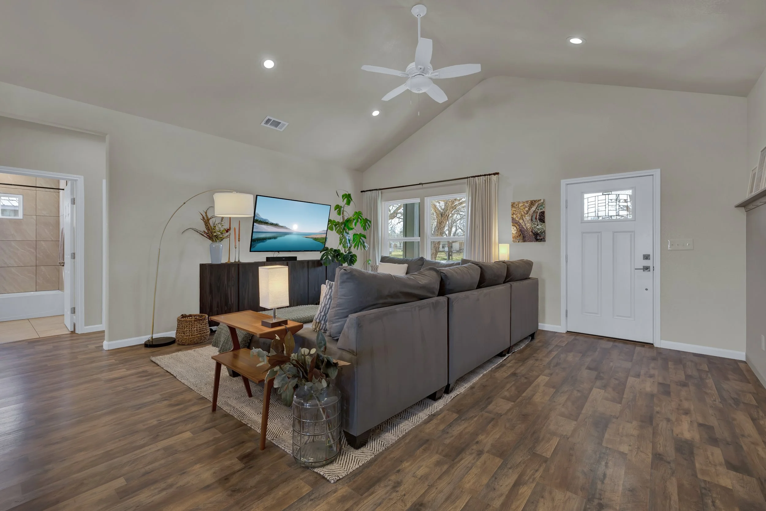 Living room with gray sectional sofa, wooden coffee table, television, large windows, and ceiling fan.