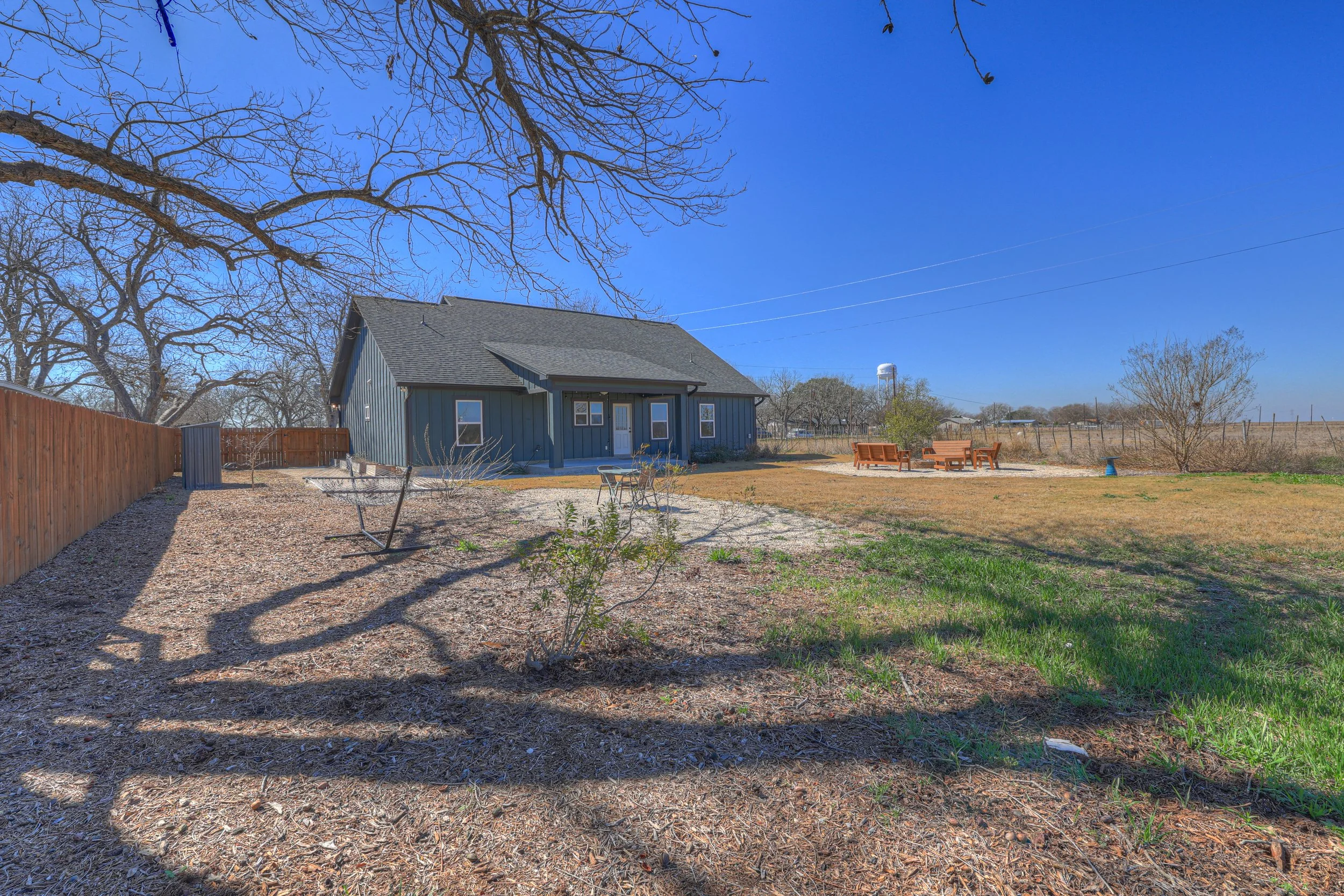 Large backyard with a blue house, leafless trees, wooden outdoor furniture, green grass patches, and a clear blue sky.