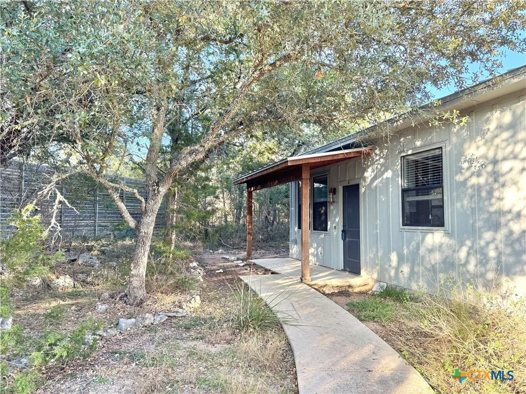 Exterior of a house with a curved concrete pathway leading to the front door, surrounded by sparse grass, shrubs, and a large tree, with a weathered metal fence in the background.