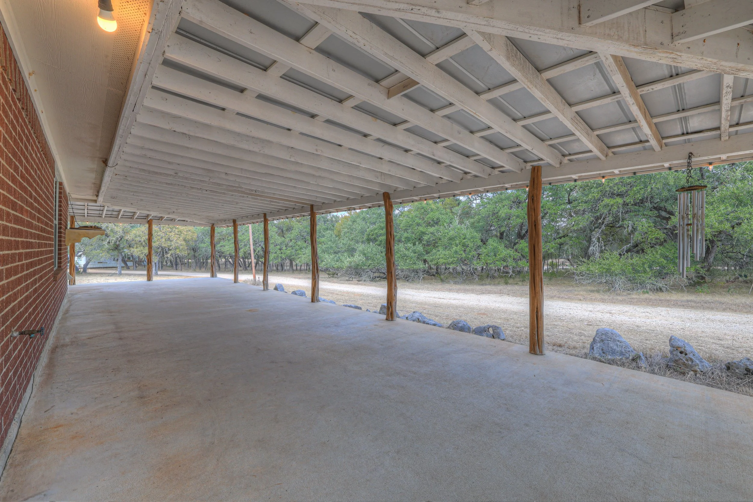 Covered patio with concrete floor, supporting wooden posts, brick wall on the left, and a wooded landscape with trees in the background.