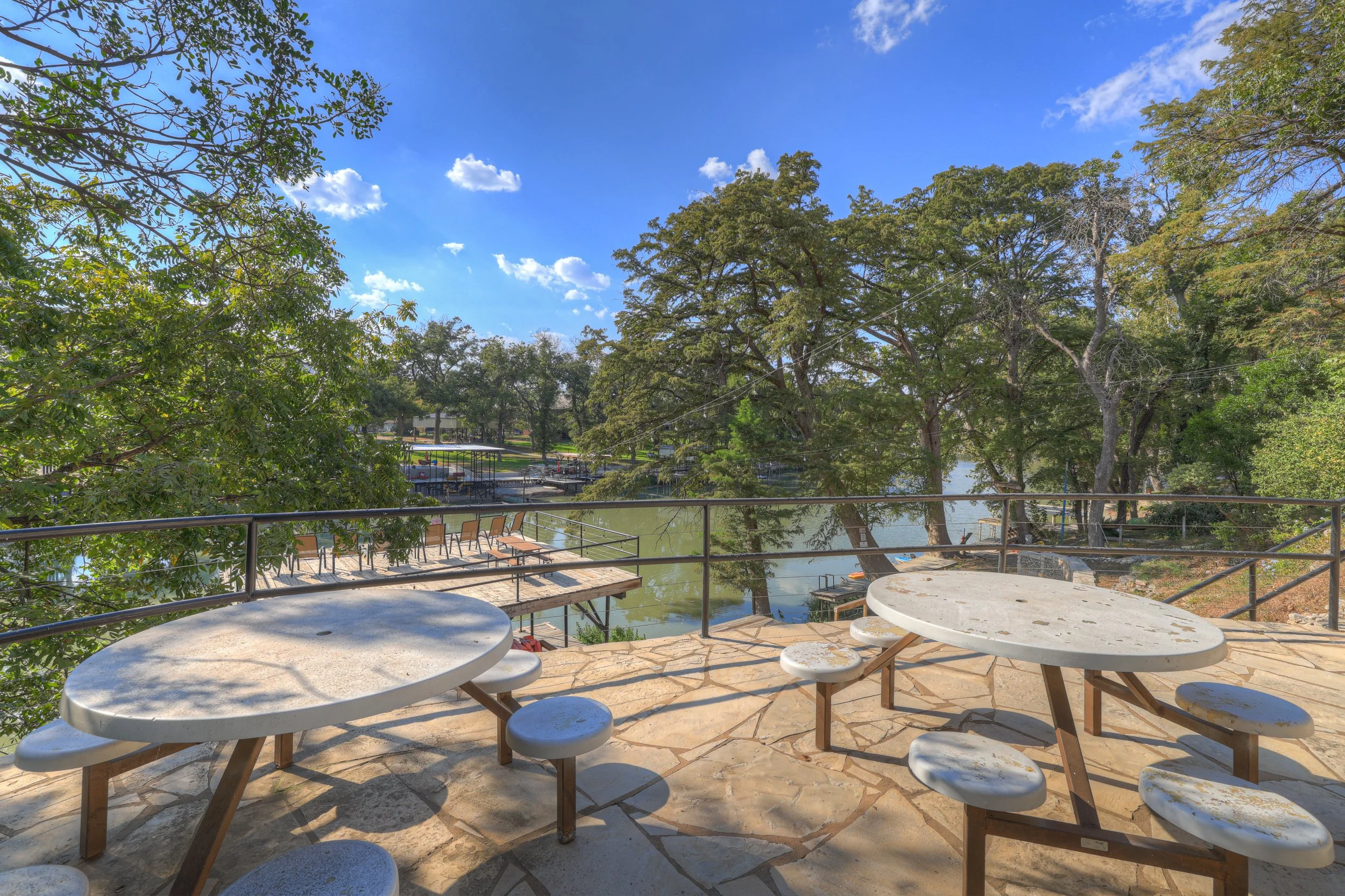Outdoor seating area with white stone tables and stools on a stone patio, overlooking a river with trees and boats, under a blue sky with scattered clouds.