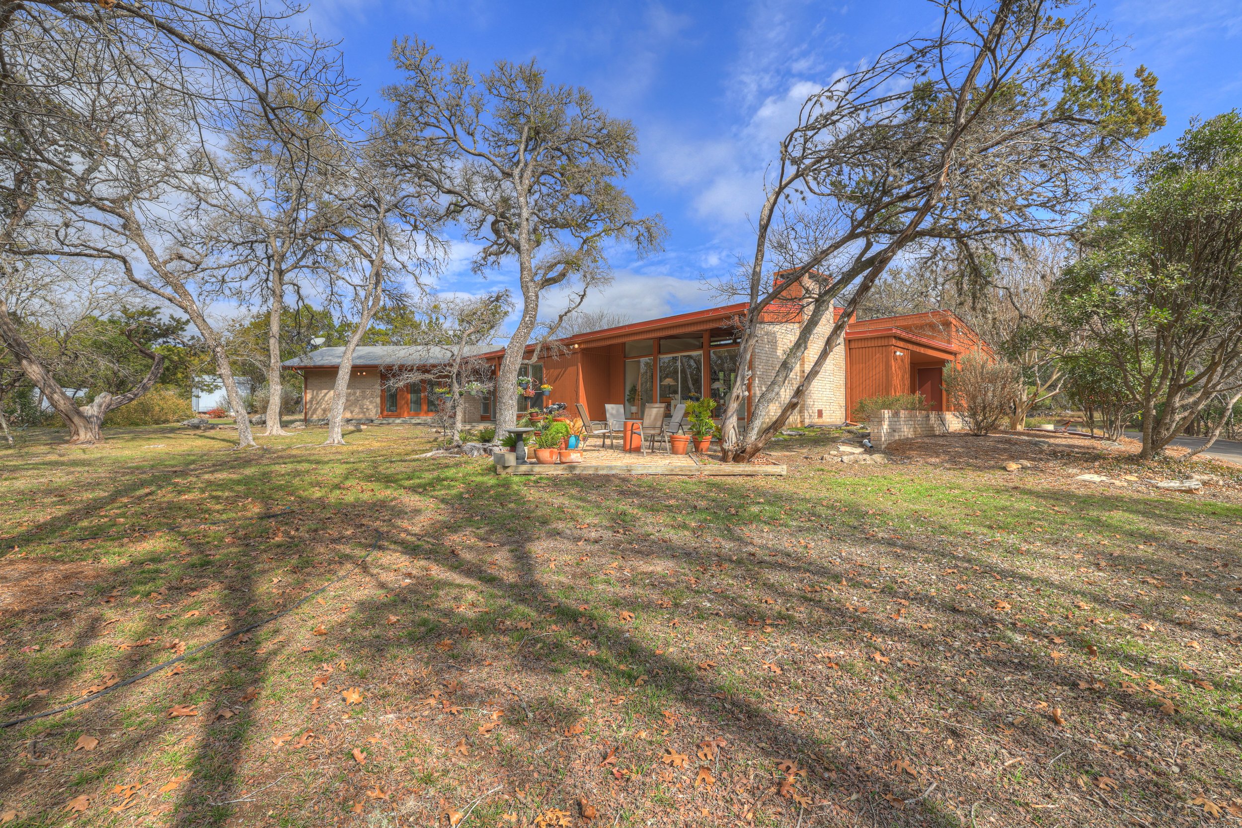 A mid-century modern house with a wooden exterior, large glass sliding doors, surrounded by leafless and green trees, and a yard with patio furniture and potted plants under a partly cloudy sky.