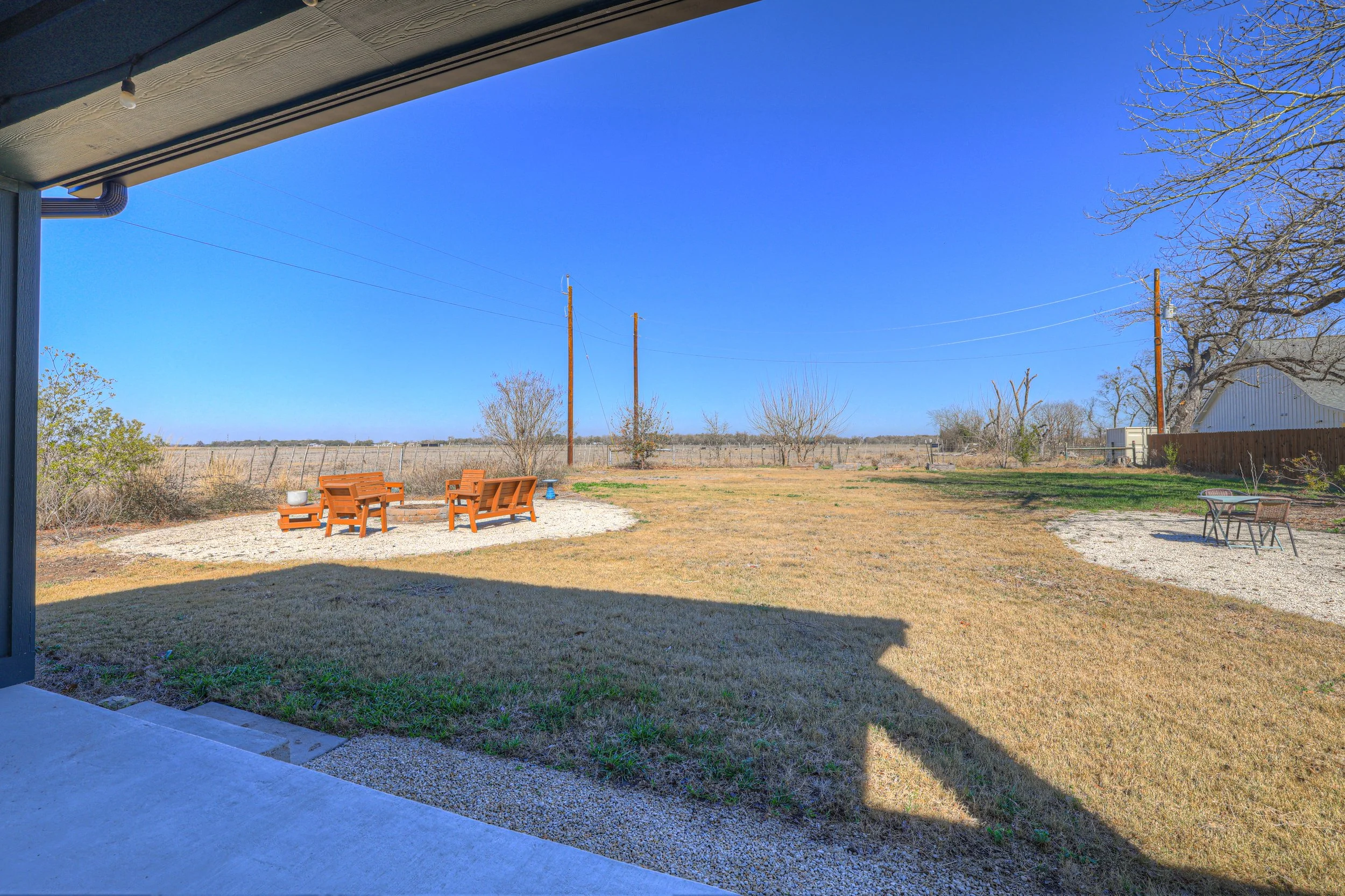 View of a backyard with a grassy area, a small seating area with wooden chairs and a table, and a larger seating area with metal chairs, under a clear blue sky with some trees and utility poles in the distance.