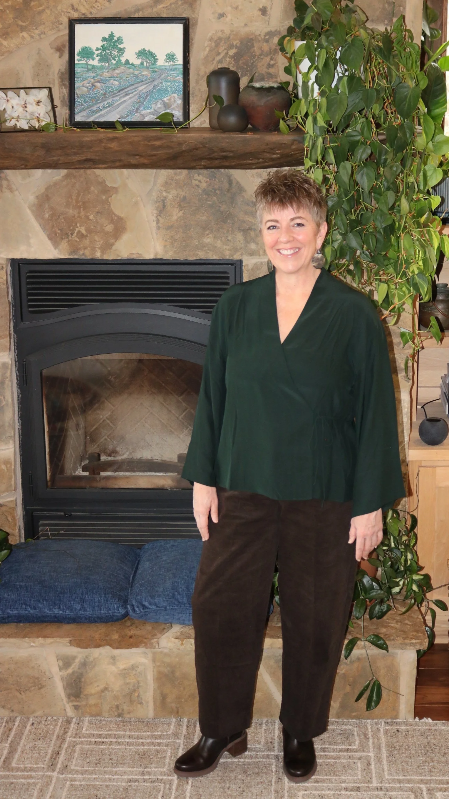 A woman standing in front of a fireplace with a stone surround, decorated with a large green plant, a picture frame, and pottery on the mantel. She is wearing a dark green top, brown pants, and black boots, smiling at the camera.