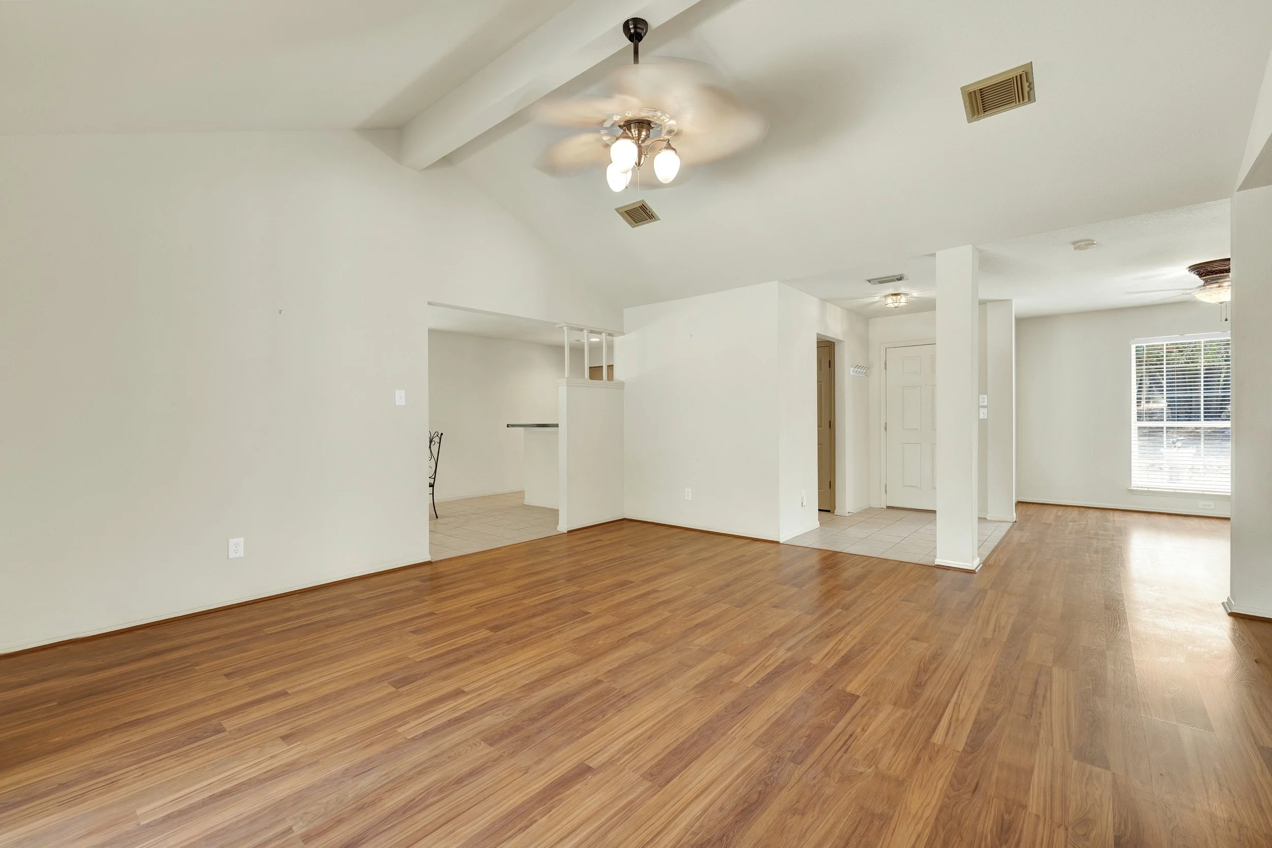 Empty living room with hardwood floors, white walls, ceiling fans, and large windows letting in natural light.