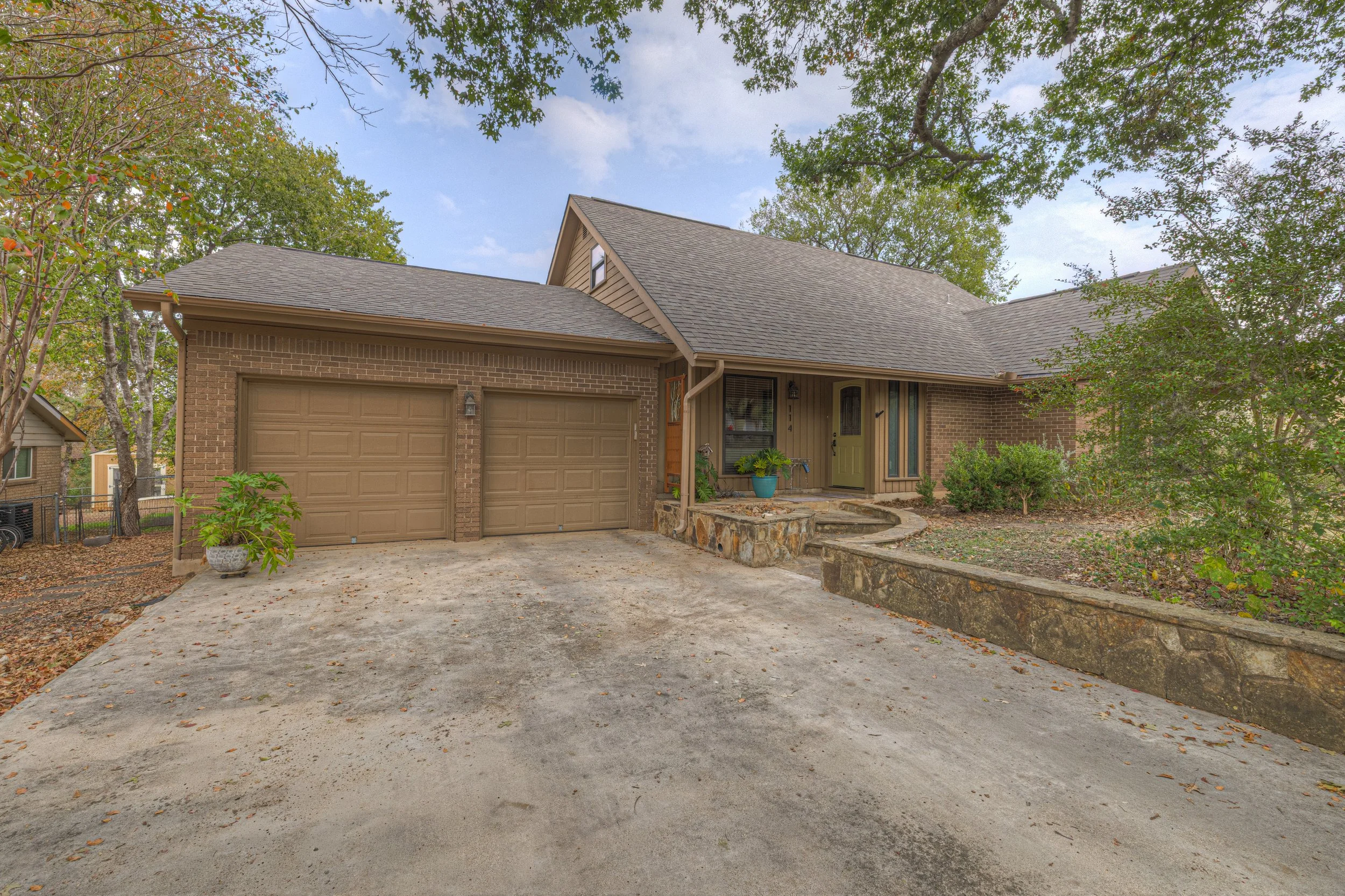 Front view of a brick house with a two-car garage, a small front porch with potted plants, and a yard with trees and shrubs.