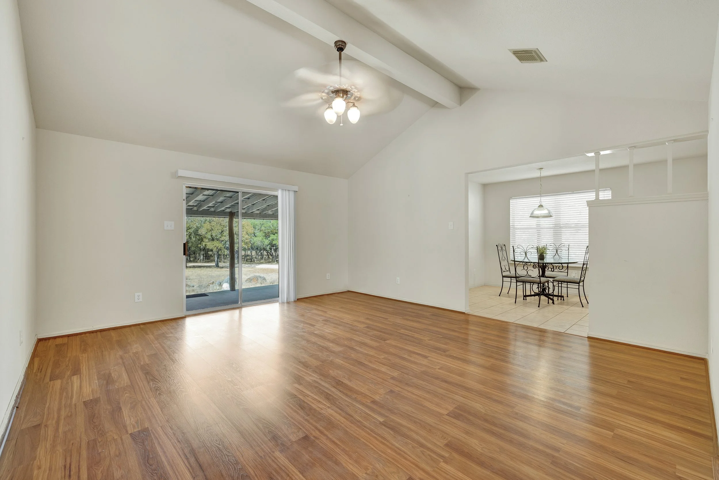 Empty living room with hardwood floors, sliding glass door to outside, ceiling fan, and opening to dining area with table and chairs.