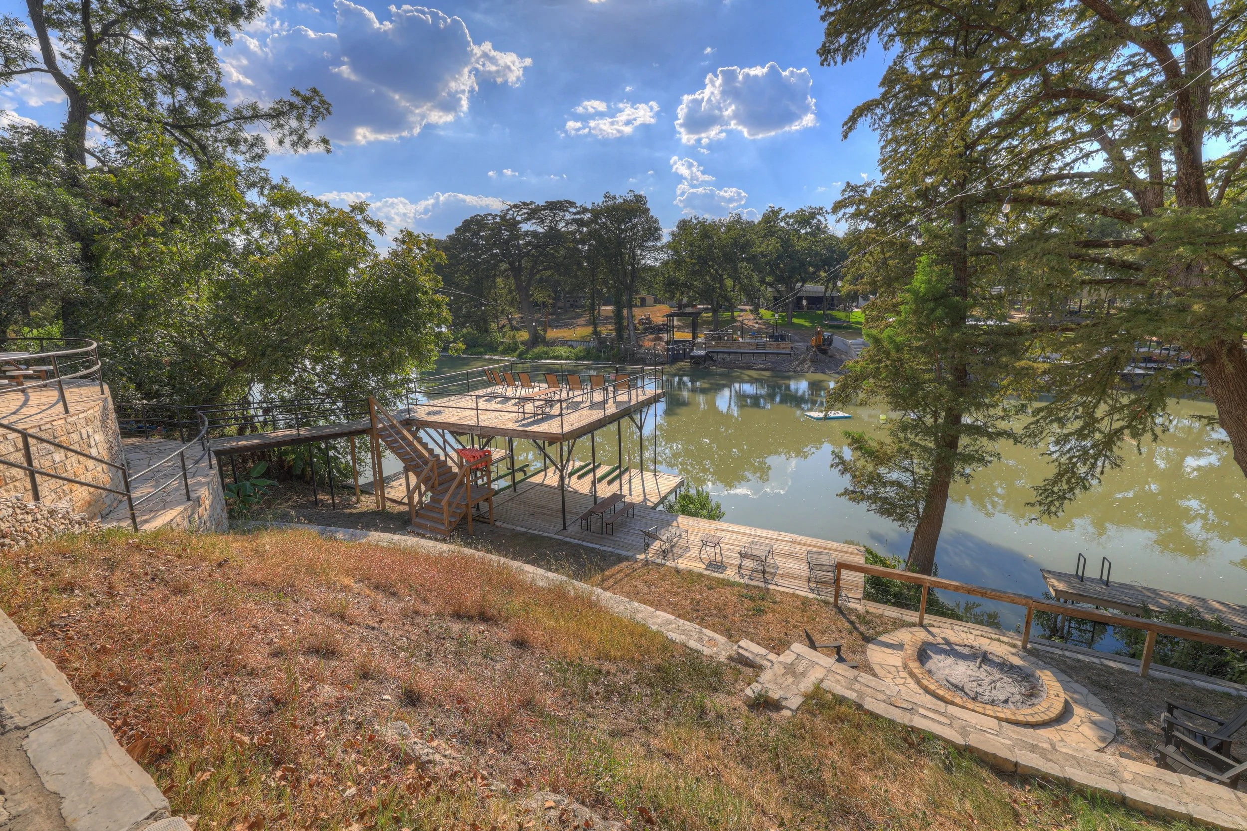 Schoolyard with a lake, trees, outdoor seating, and a fire pit, under a partly cloudy sky.