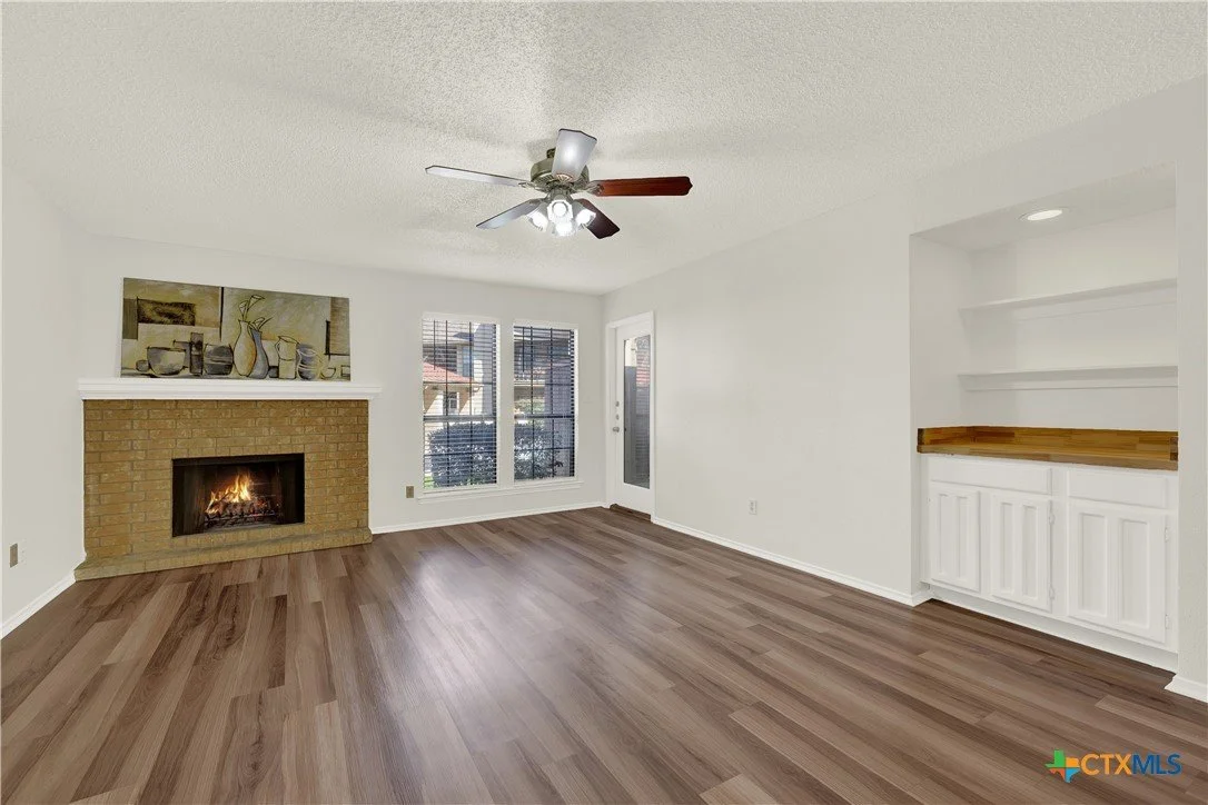 Living room with white walls, a brick fireplace with a fire burning, a ceiling fan with lights, hardwood flooring, three windows, a door, and built-in white shelves with cabinets.