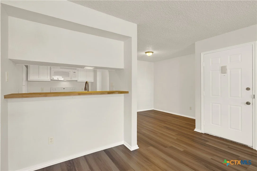 Empty living room with a small kitchen opening, white walls, wood flooring, and a front door.