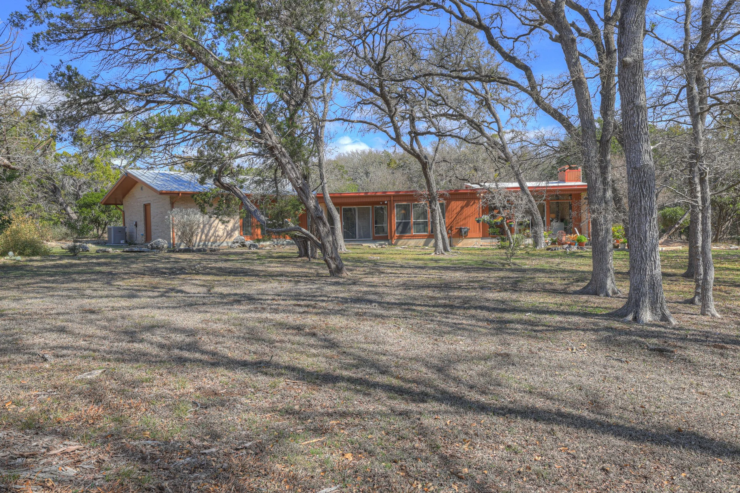 A house with a brown exterior, large windows, and a patio, surrounded by leafless trees on a partly cloudy day.
