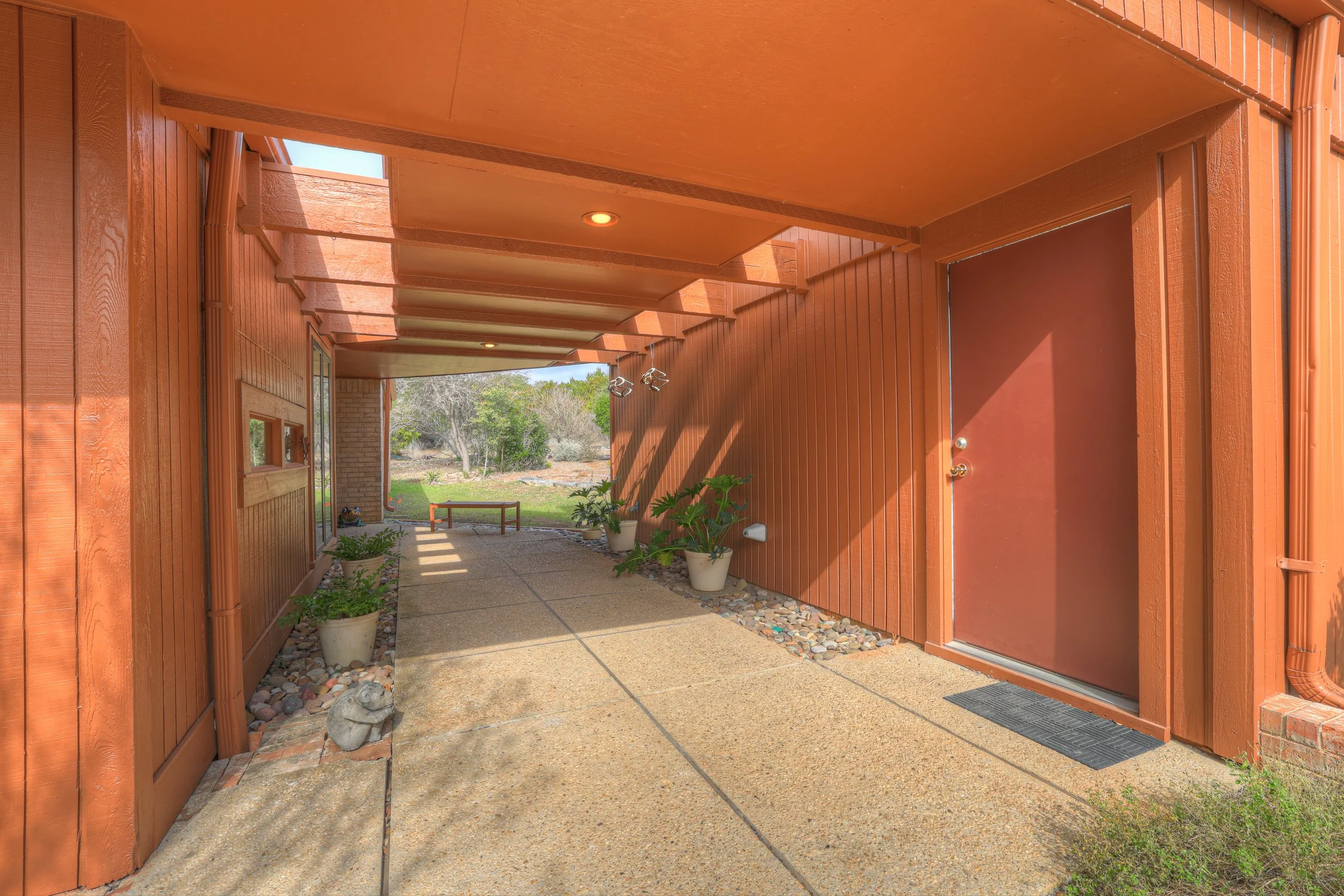 Covered outdoor walkway leading to a garden area, with potted plants along the side, a bench at the end, and a brown door on the right.