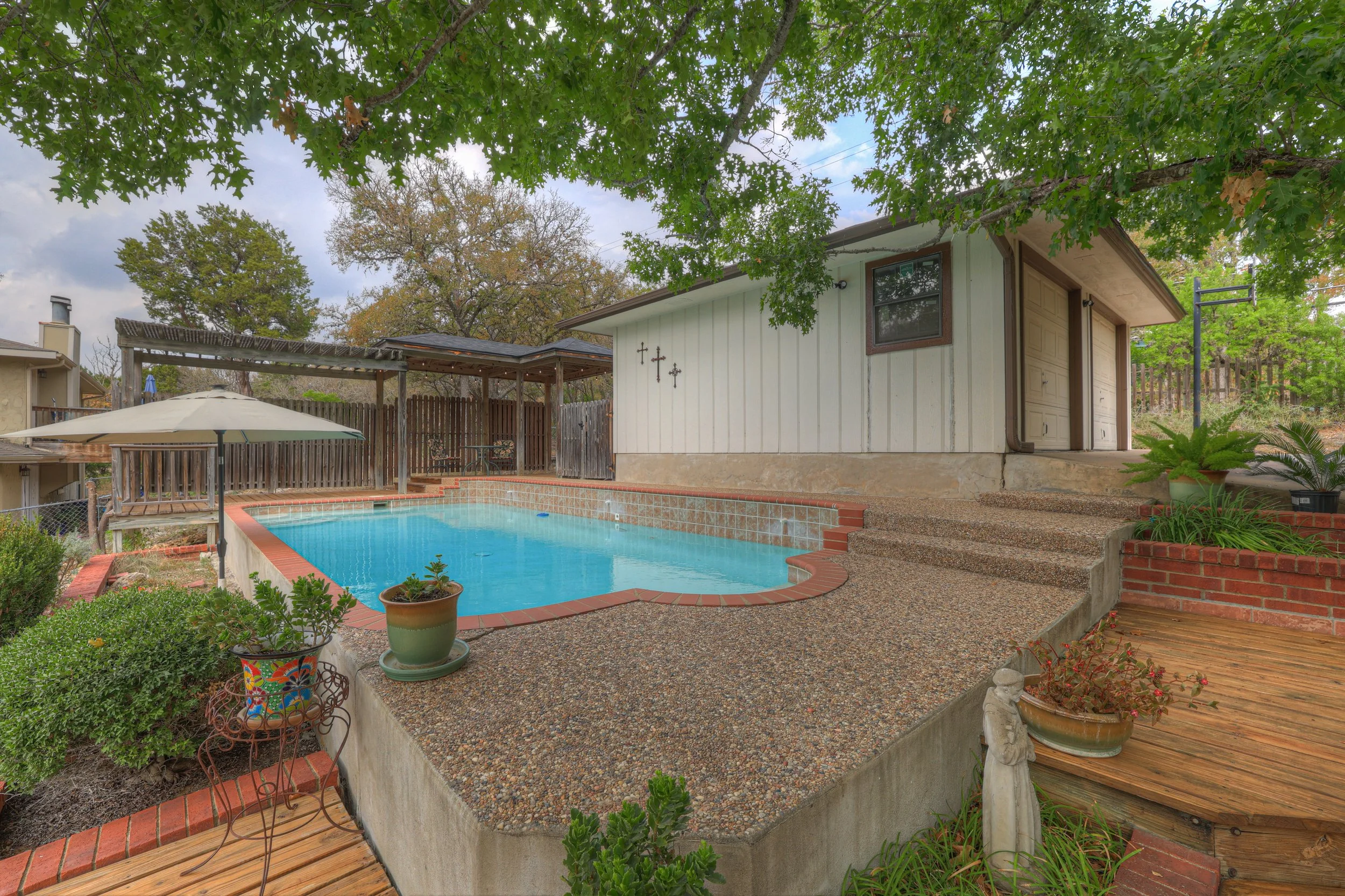 Backyard with swimming pool, potted plants, deck, umbrella, trees, and small building or shed.