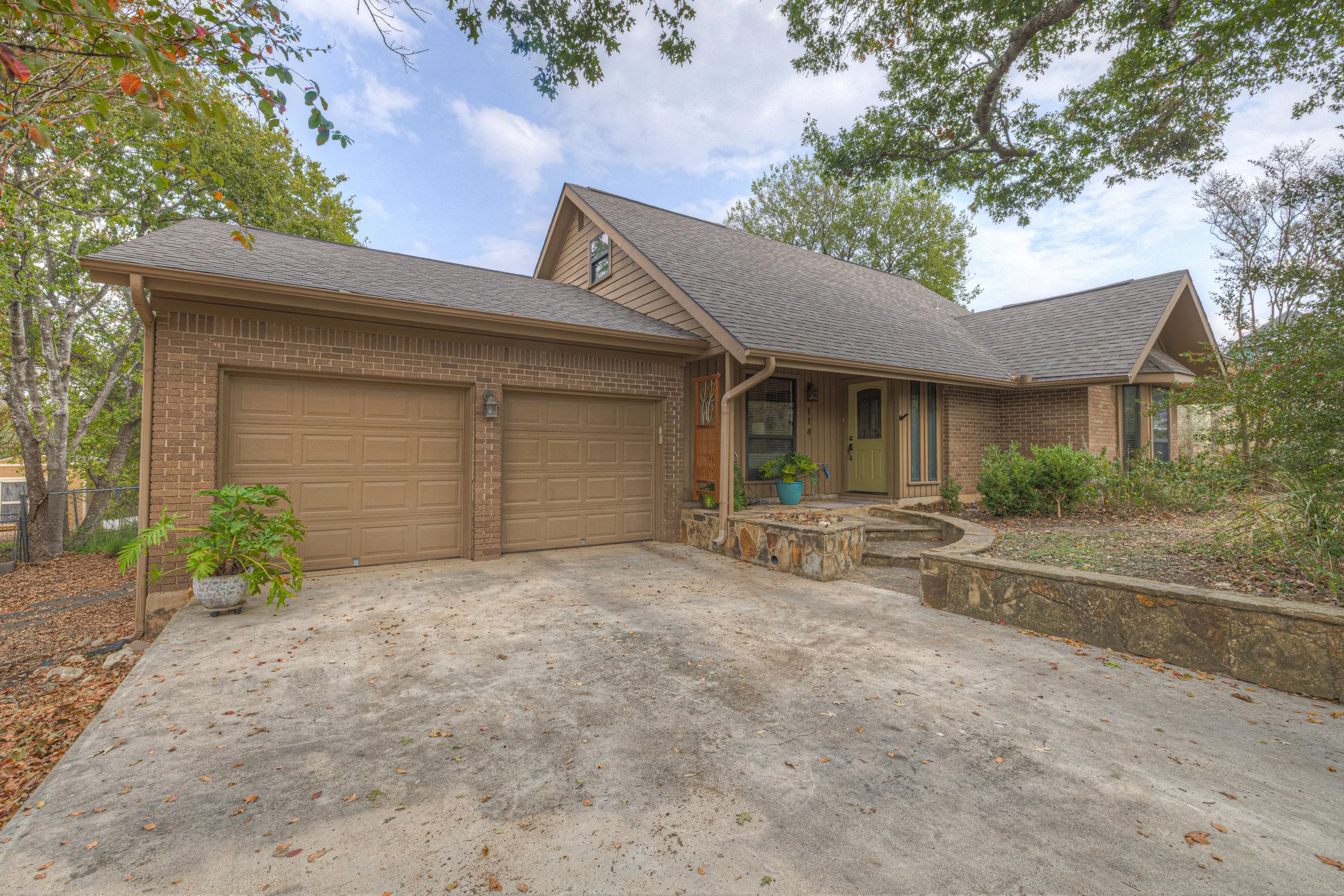Front view of a brick and siding house with a two-car garage, concrete driveway, and a small front yard with plants and trees, under a partly cloudy sky.