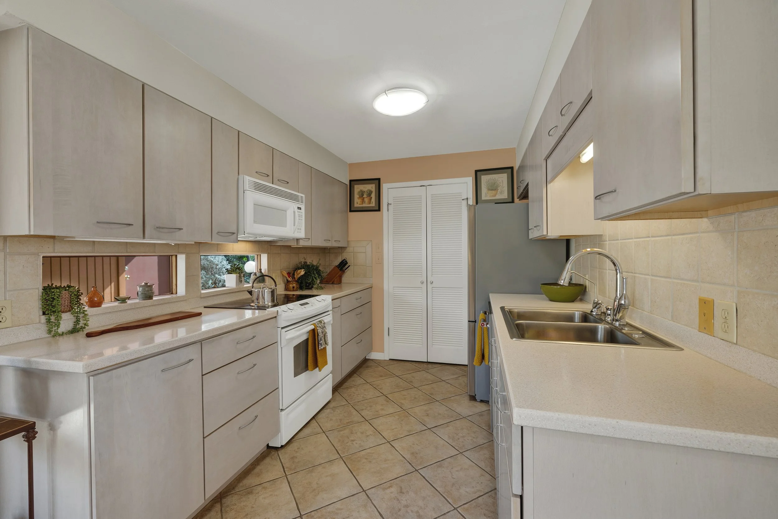 Kitchen with beige cabinets, white countertops, and appliances including a microwave, stove, and refrigerator. There are small windows above the countertop and a double sink with a green bowl.