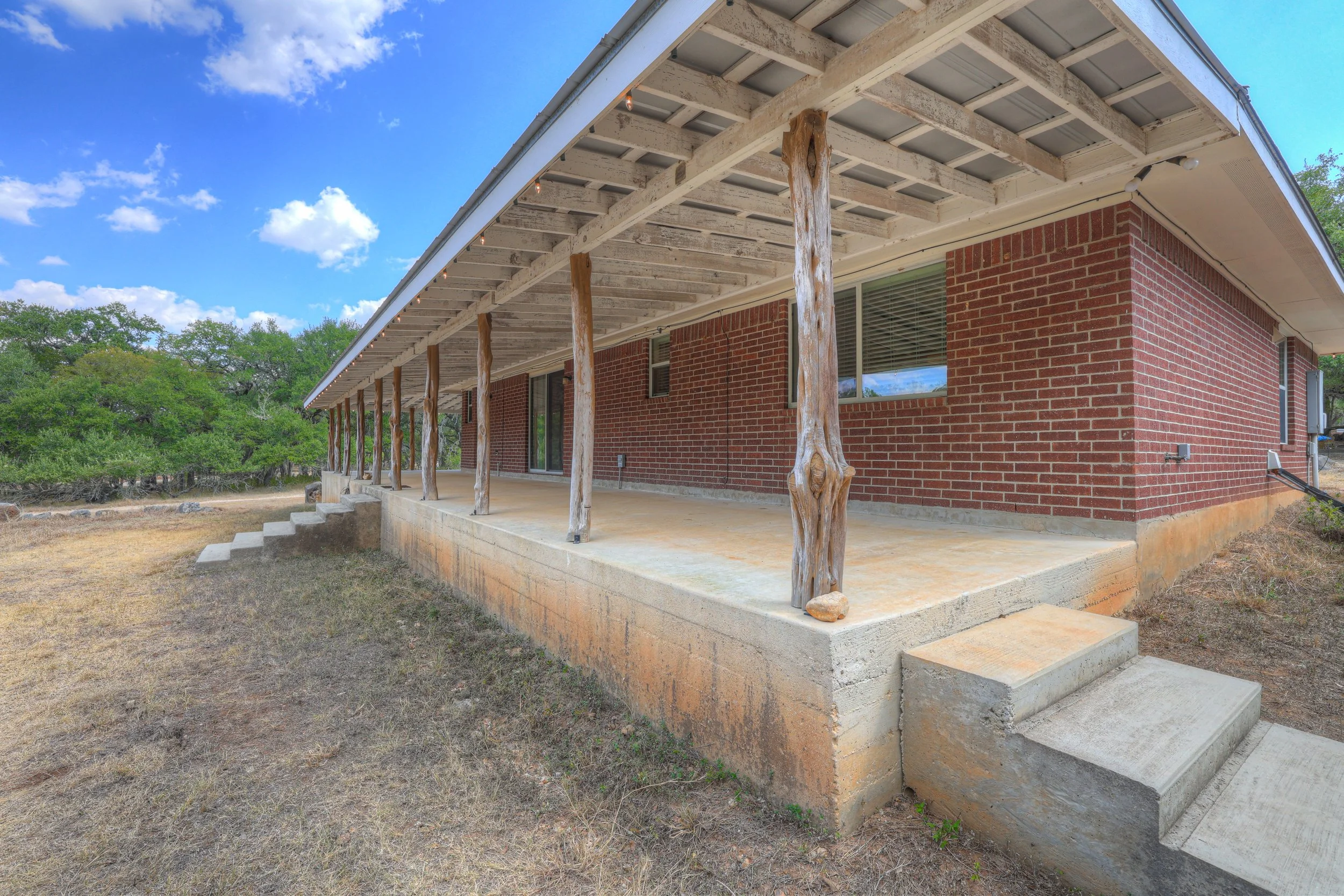 A brick house with a large, raised porch supported by uneven, rustic wooden posts, featuring a concrete foundation and steps leading up to the porch, set against a background of green trees and a blue sky with clouds.