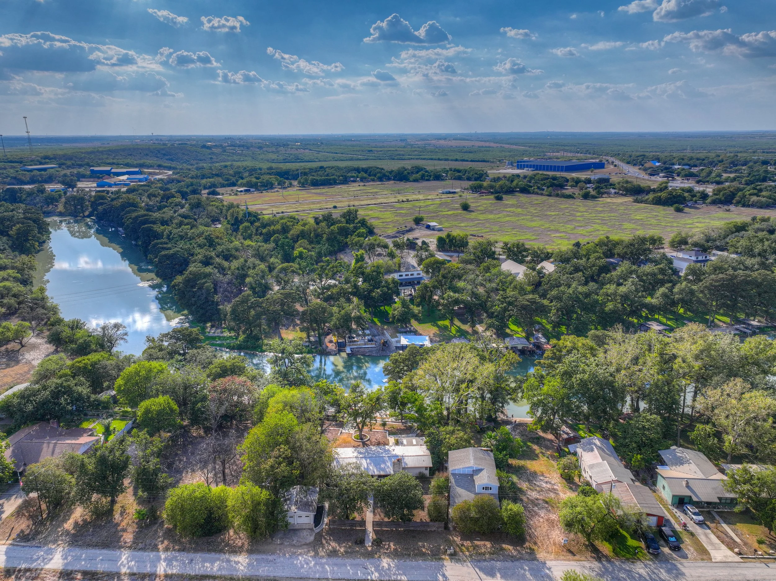 Aerial view of a river flowing through a green residential area with homes, trees, and yards, with open fields and distant buildings under a partly cloudy sky.