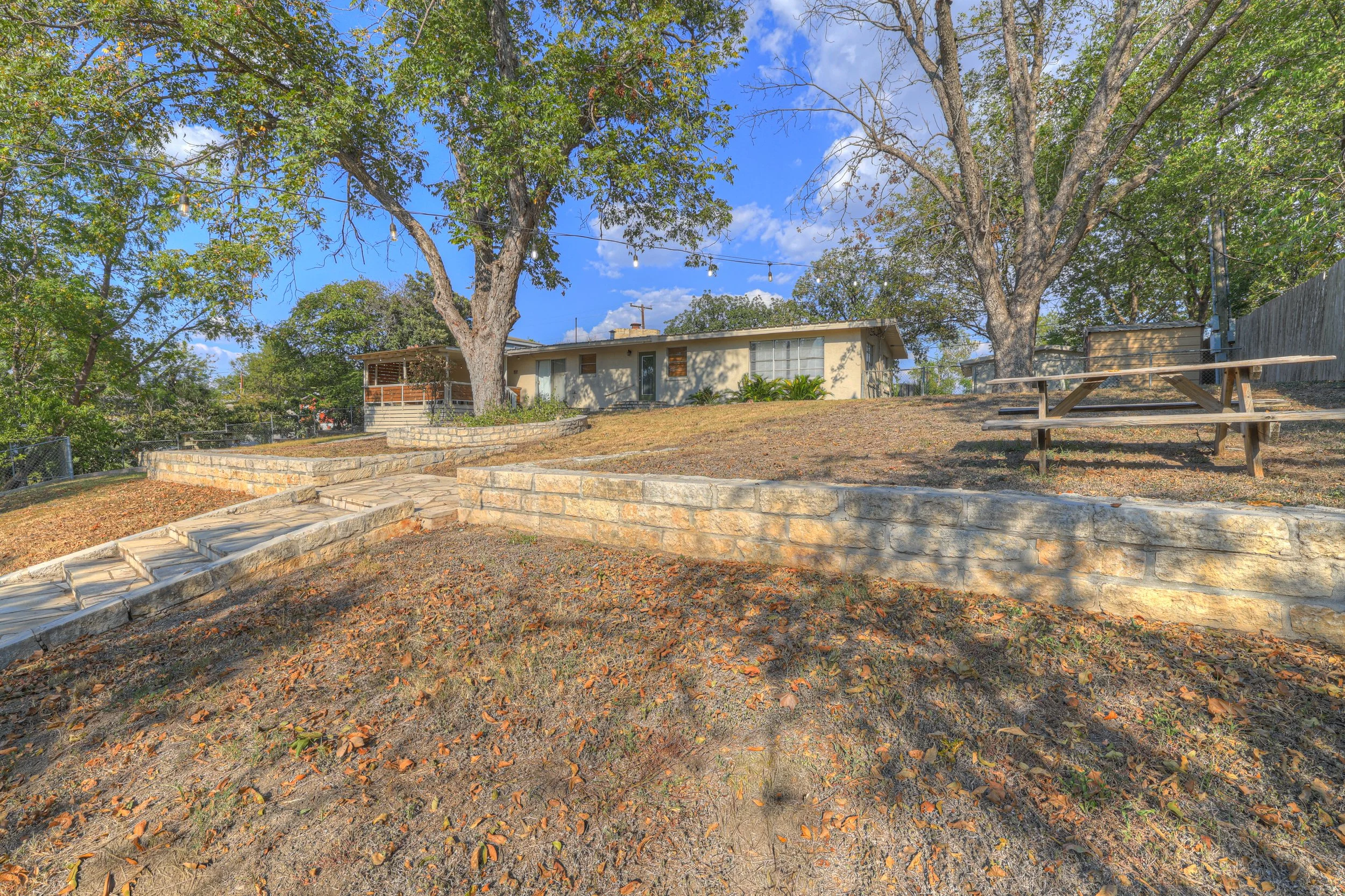 Backyard with stone steps and retaining walls, trees providing shade, picnic table, and a house in the background under a blue sky with scattered clouds.
