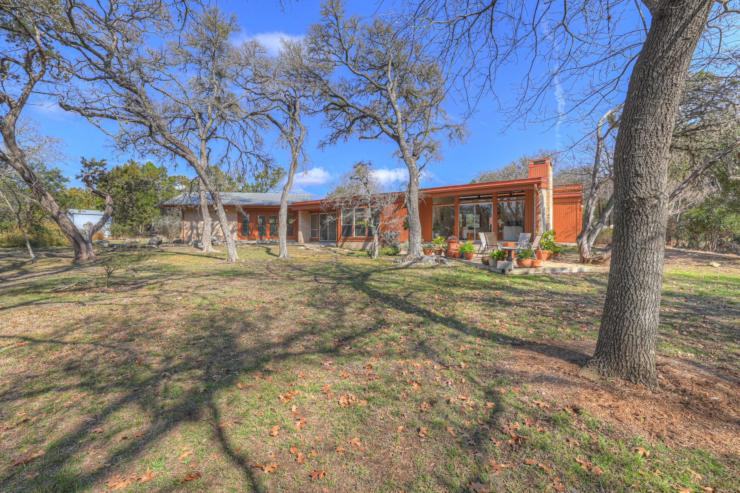 A single-story house with a large wooden deck, surrounded by leafless trees and a grassy yard on a sunny day.