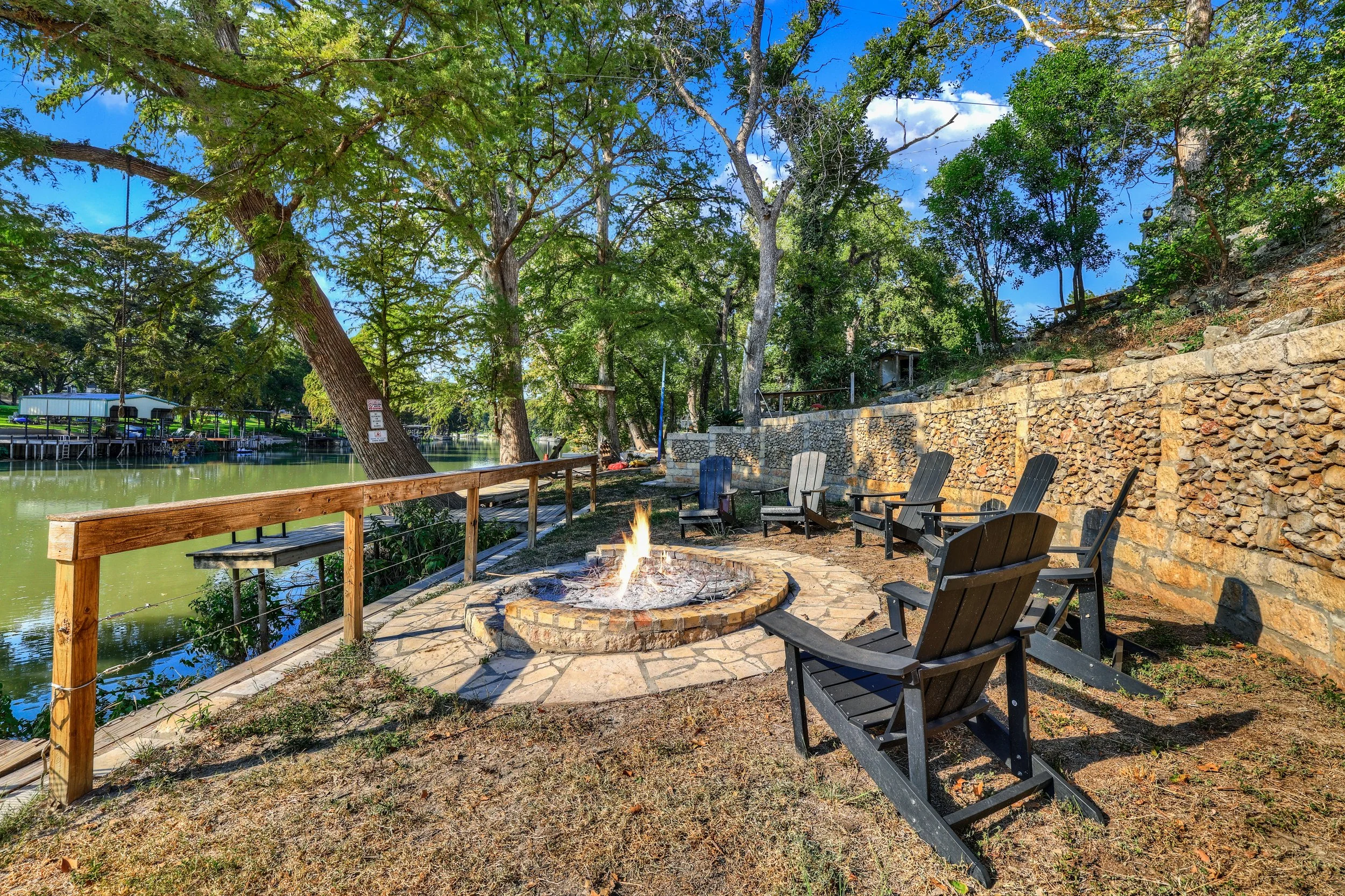 Seating area with Adirondack chairs around a fire pit by a lake, with trees and a stone retaining wall in the background.