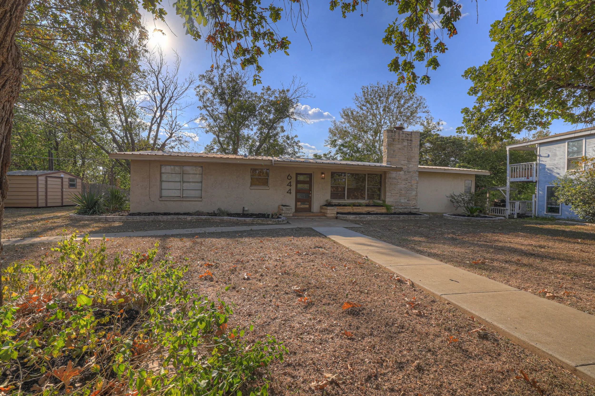 Single-story house with beige exterior and stone chimney, front yard with a concrete walkway, and surrounded by sparse trees and bushes on a sunny day.
