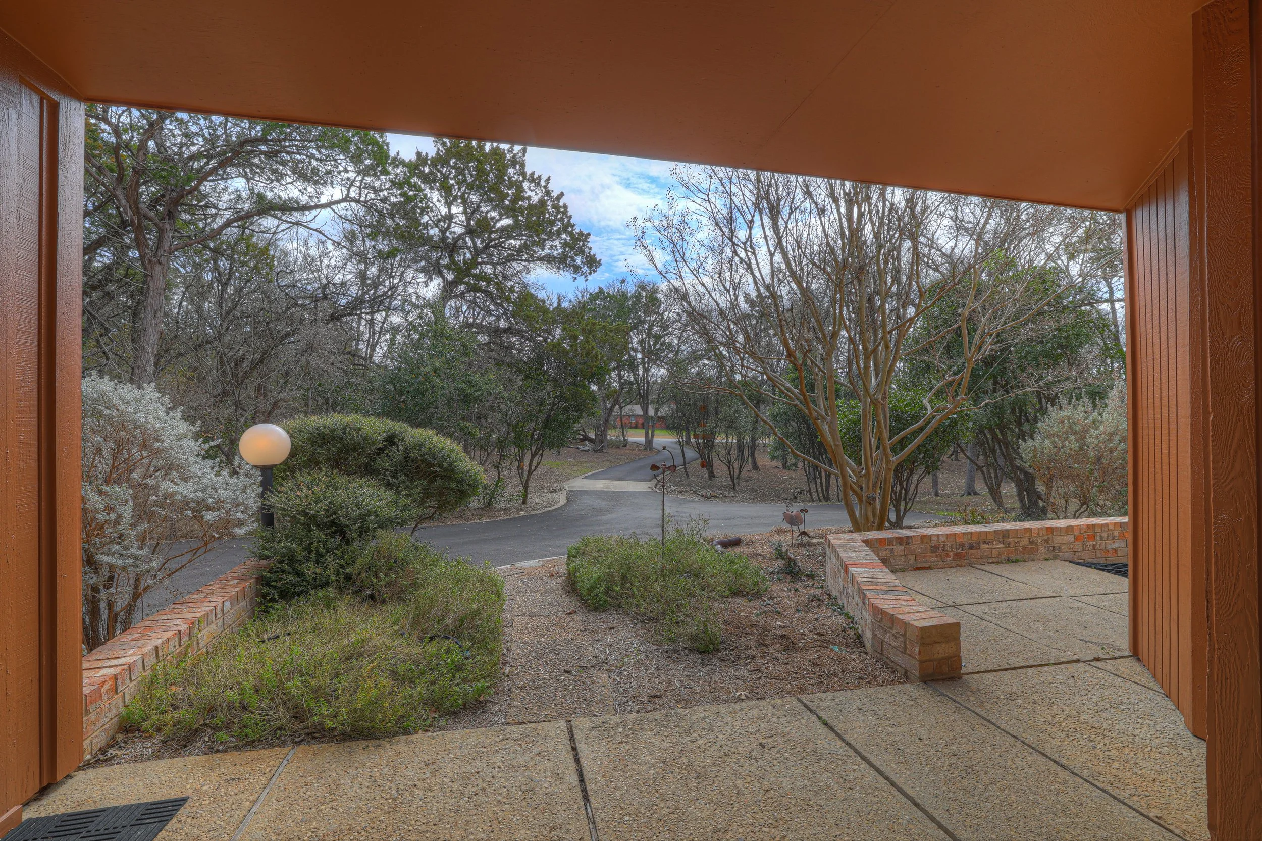 View from a porch or patio area looking out onto a landscaped yard with trees, bushes, a winding pathway, and decorative garden art, under a partly cloudy sky.
