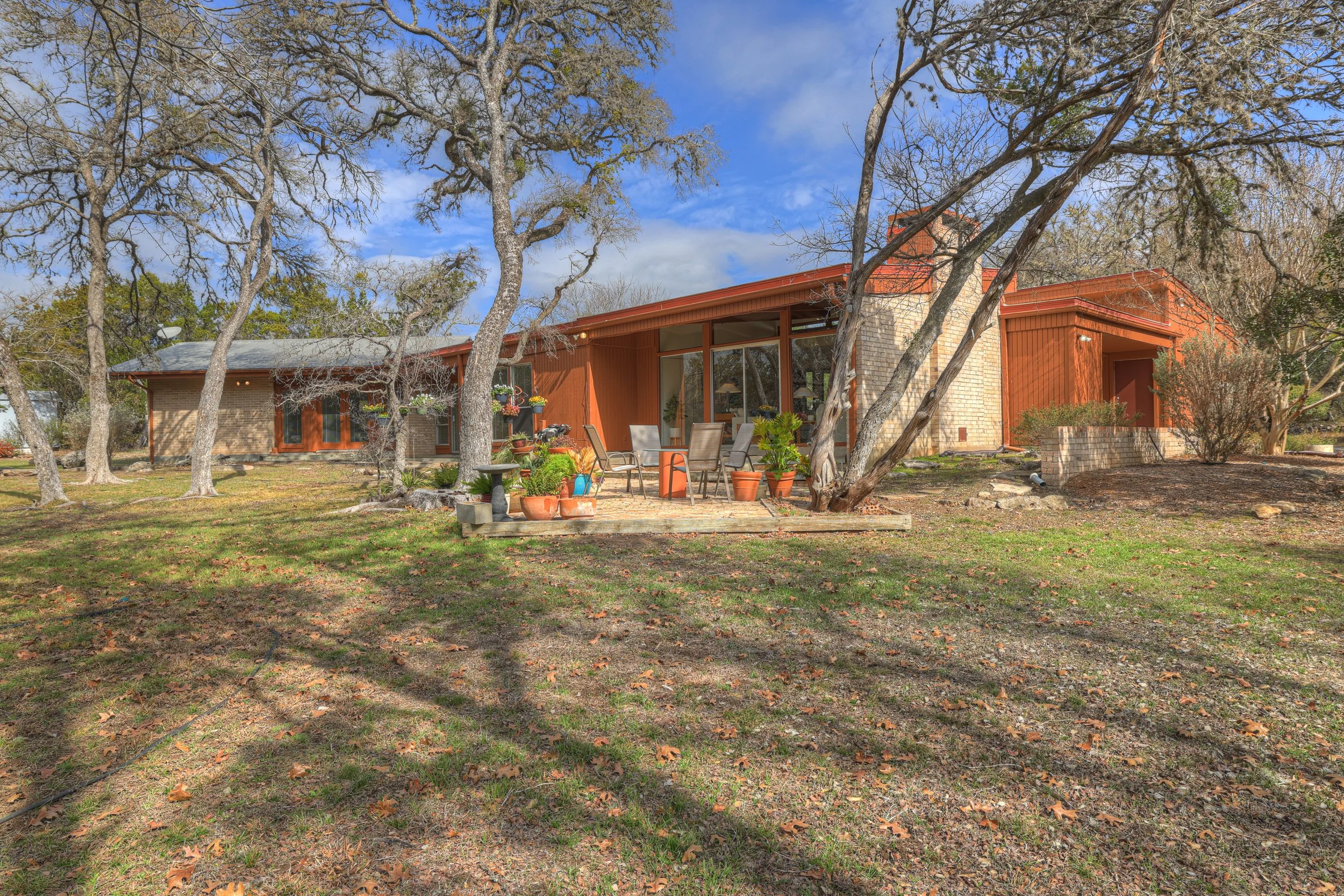 A mid-century modern house with a large glass sliding door, situated in a yard with leafless trees and a small patio area with outdoor furniture and potted plants.