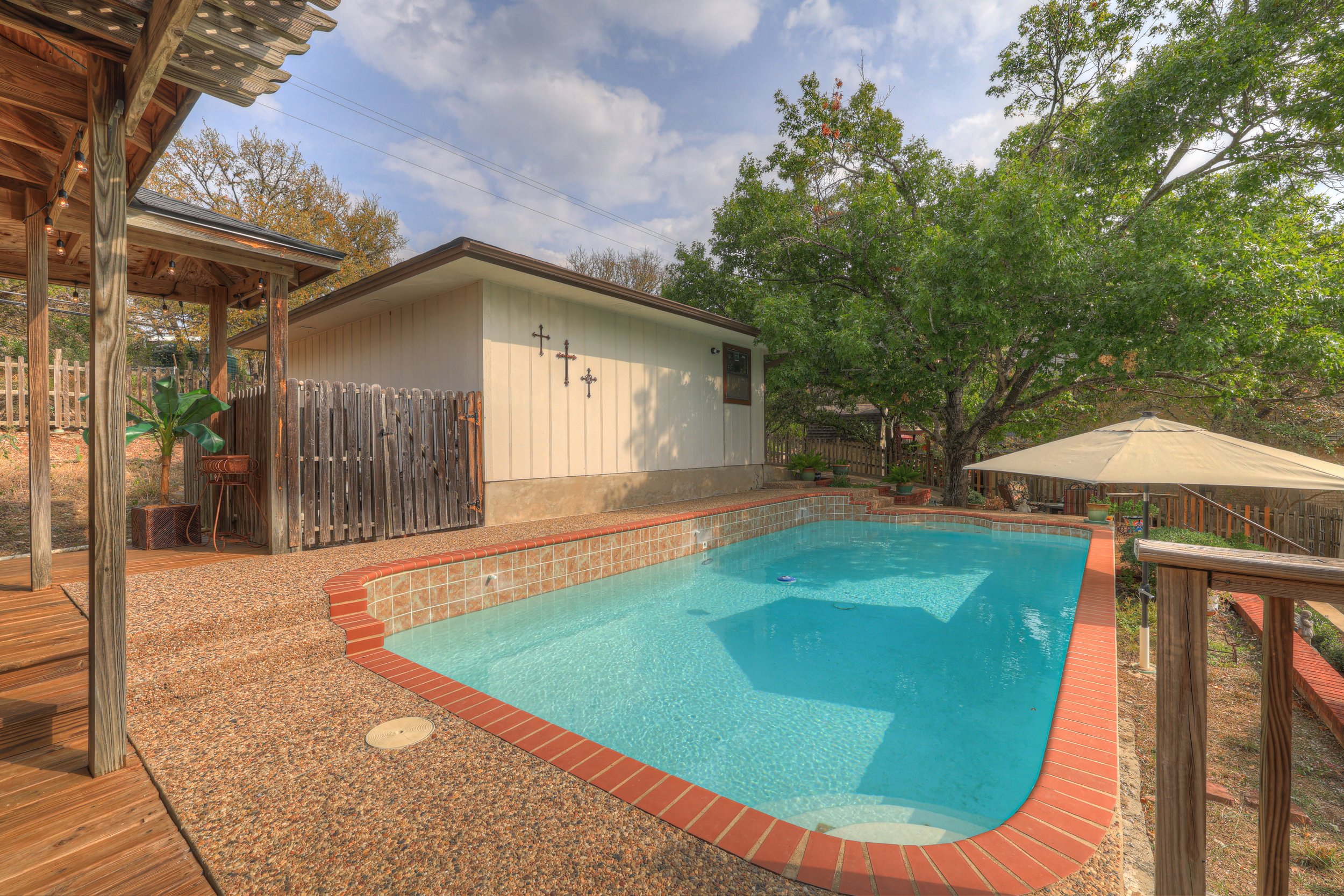 A backyard with a swimming pool, umbrella, large tree, and a small building with Cross decorations on the wall.