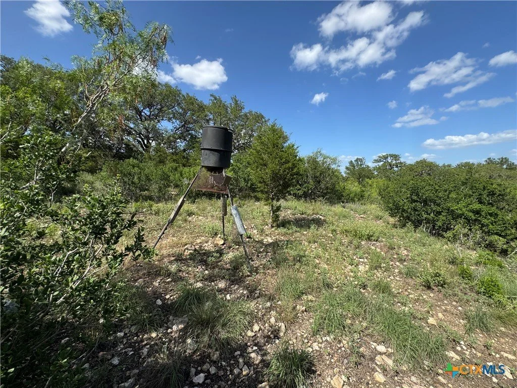 A windmill with a black top and a mesh base standing in a grassy, rocky field with sparse trees and bushes under a blue sky with scattered clouds.