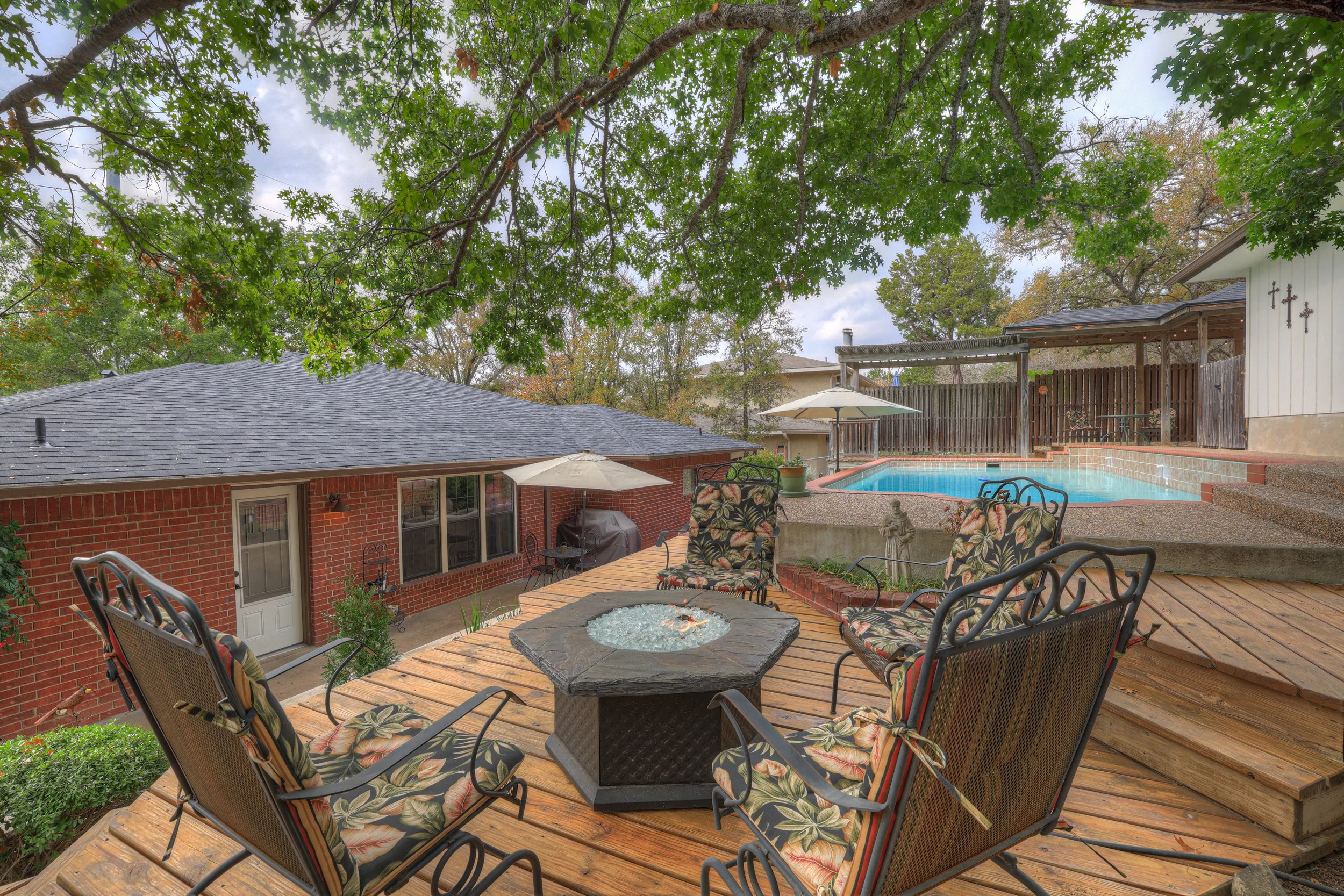 Backyard patio with wooden seating area, firepit, umbrellas, potted plants, and a swimming pool enclosed by a wooden fence, surrounded by green trees.