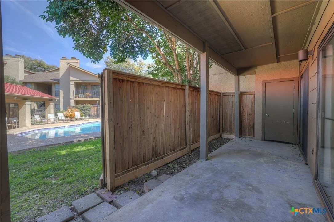 Private patio area with a concrete floor, wooden fence, and a view of a swimming pool and outdoor seating at an apartment complex.